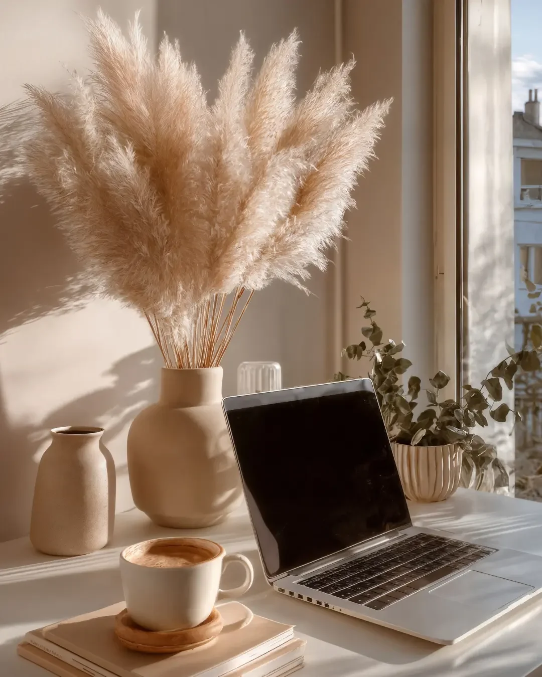 A minimalistic workspace with a laptop, a white cup of coffee on a saucer, a stack of papers or notebooks, and decorative vases with dried pampas grass and greenery by a window with soft sunlight.