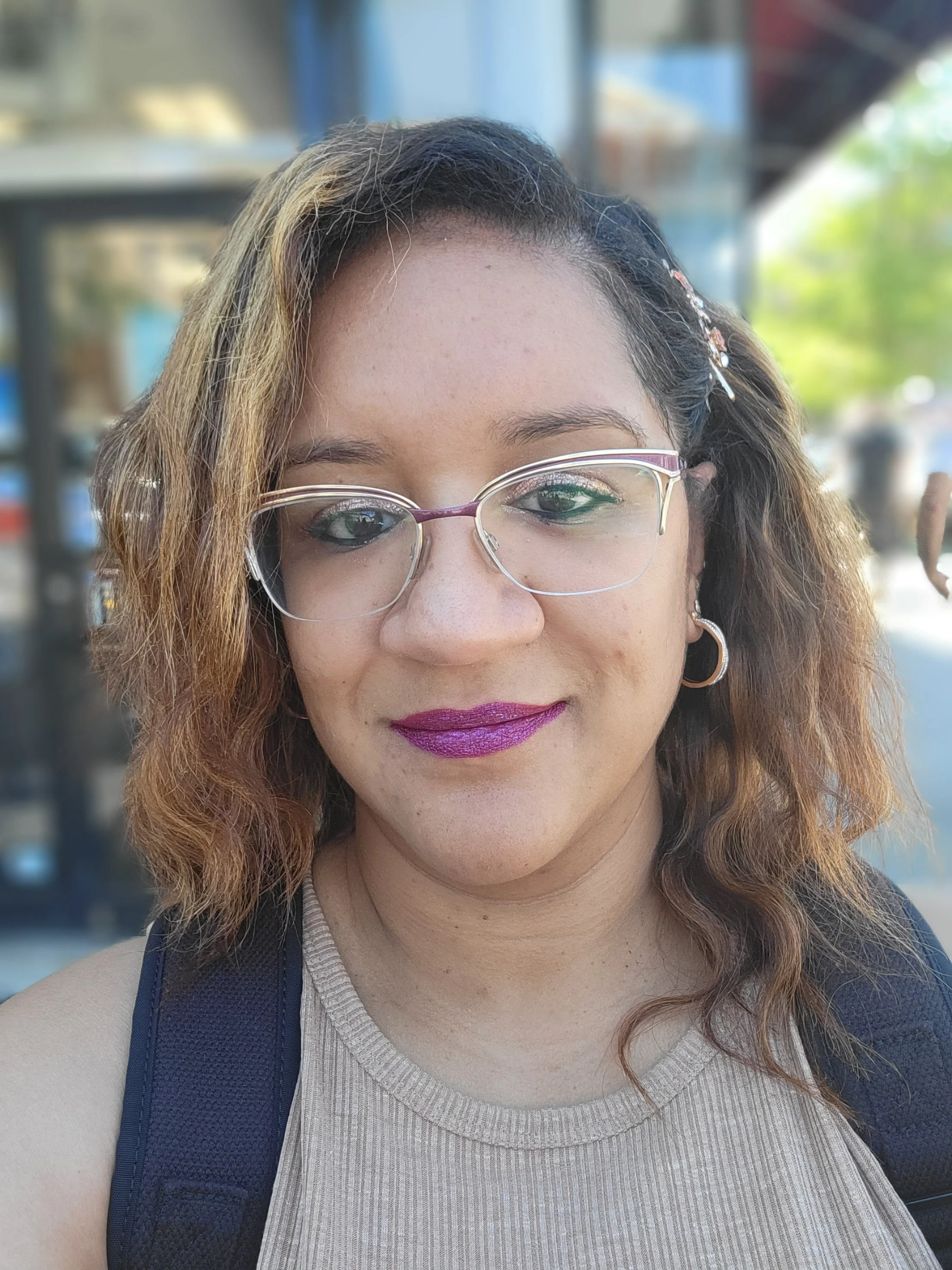 A woman with light skin, wavy hair with darker roots and blonde highlights, wearing glasses, purple lipstick, and hoop earrings, wearing a beige sleeveless top, standing outdoors with blurred background of trees and buildings.