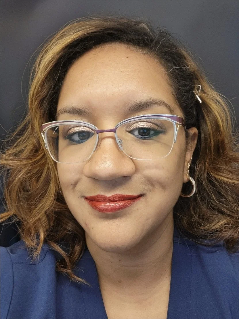A woman with wavy, shoulder-length hair, glasses, red lipstick, and a dark blue shirt, smiling at the camera