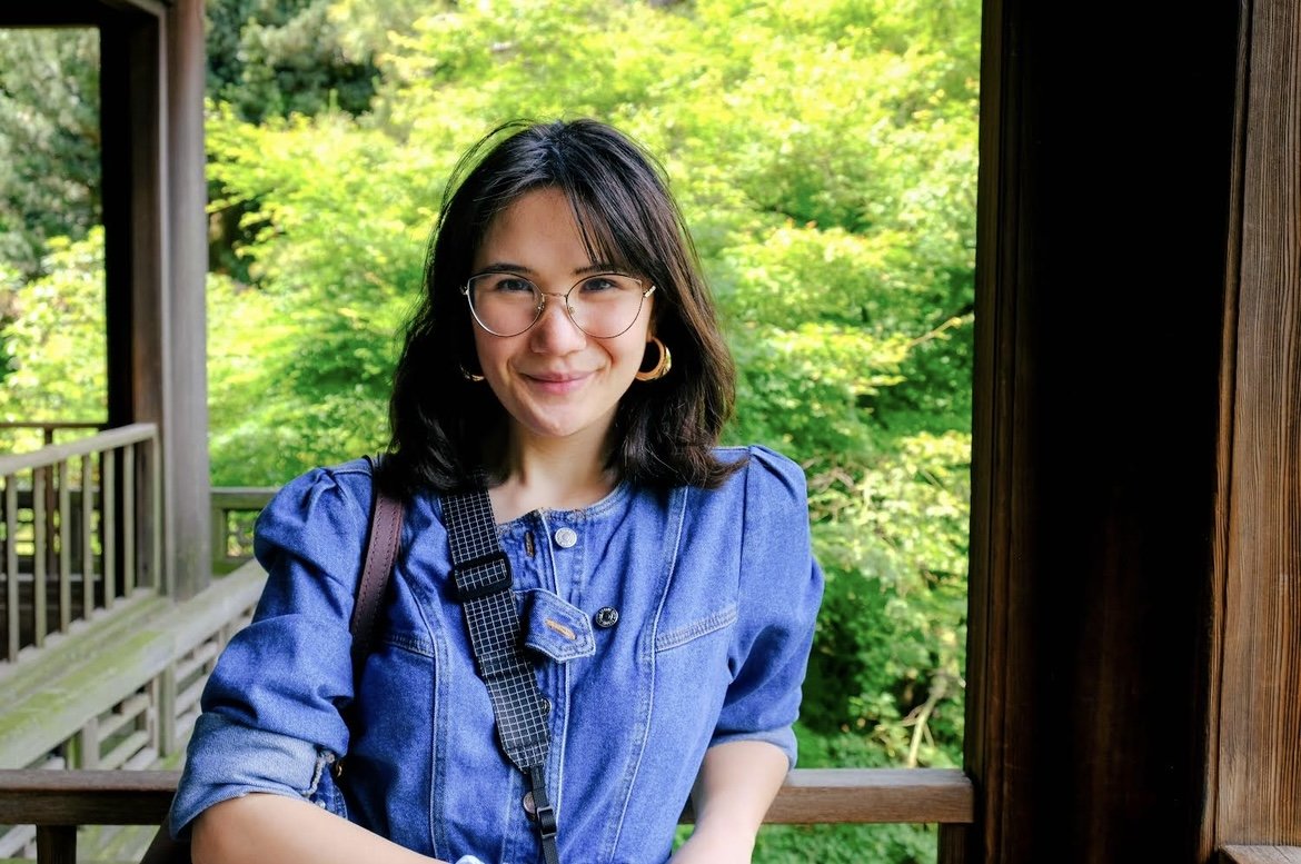 A young woman with dark hair and glasses smiling outdoors on a wooden balcony surrounded by lush green trees.