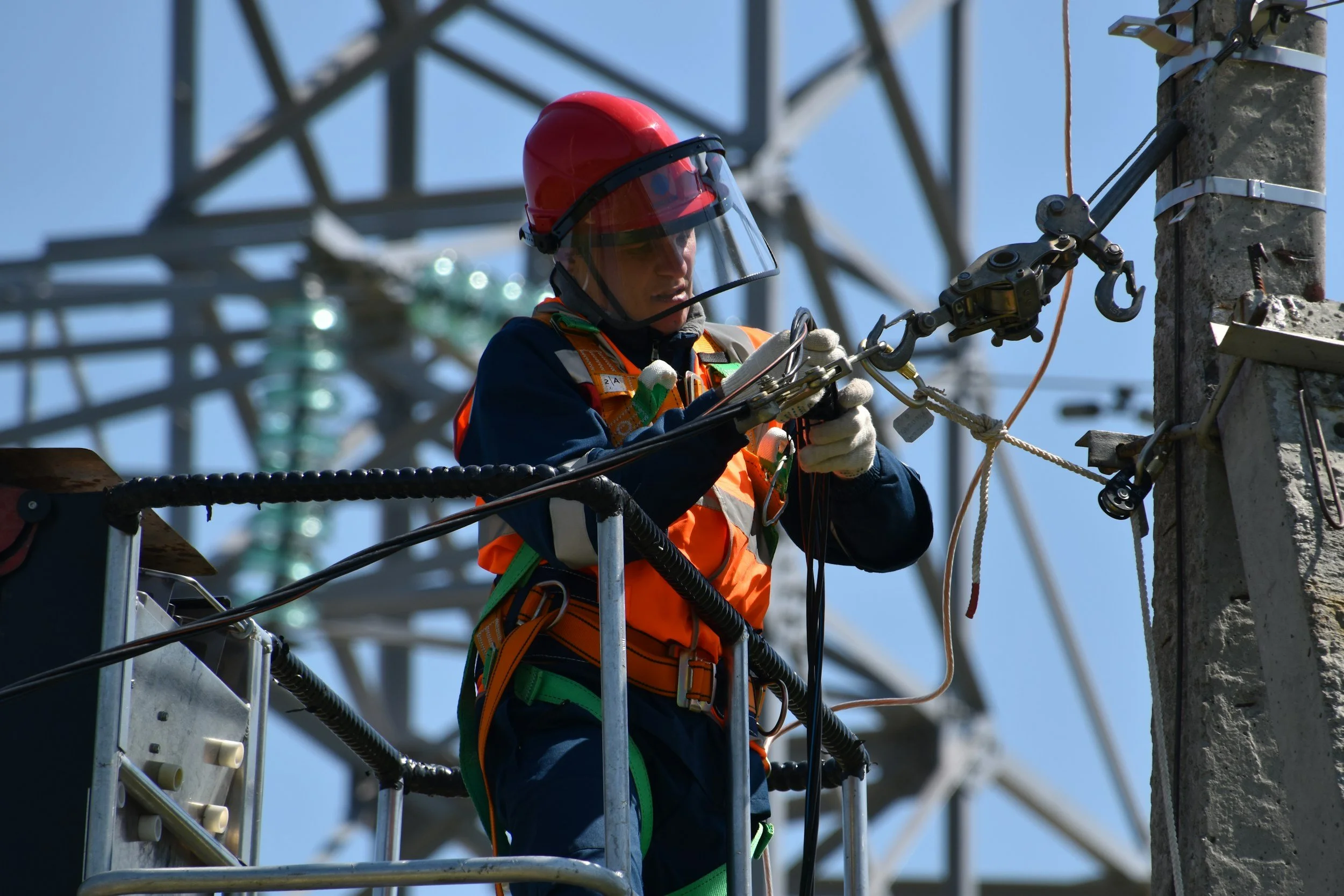 A worker wearing safety gear, including a red helmet and safety harness, working on power lines on an electrical tower.