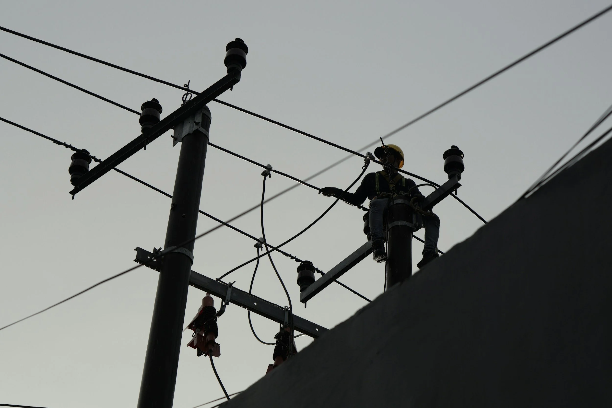 A worker wearing a safety helmet and harness working on an electrical power pole during overcast weather.