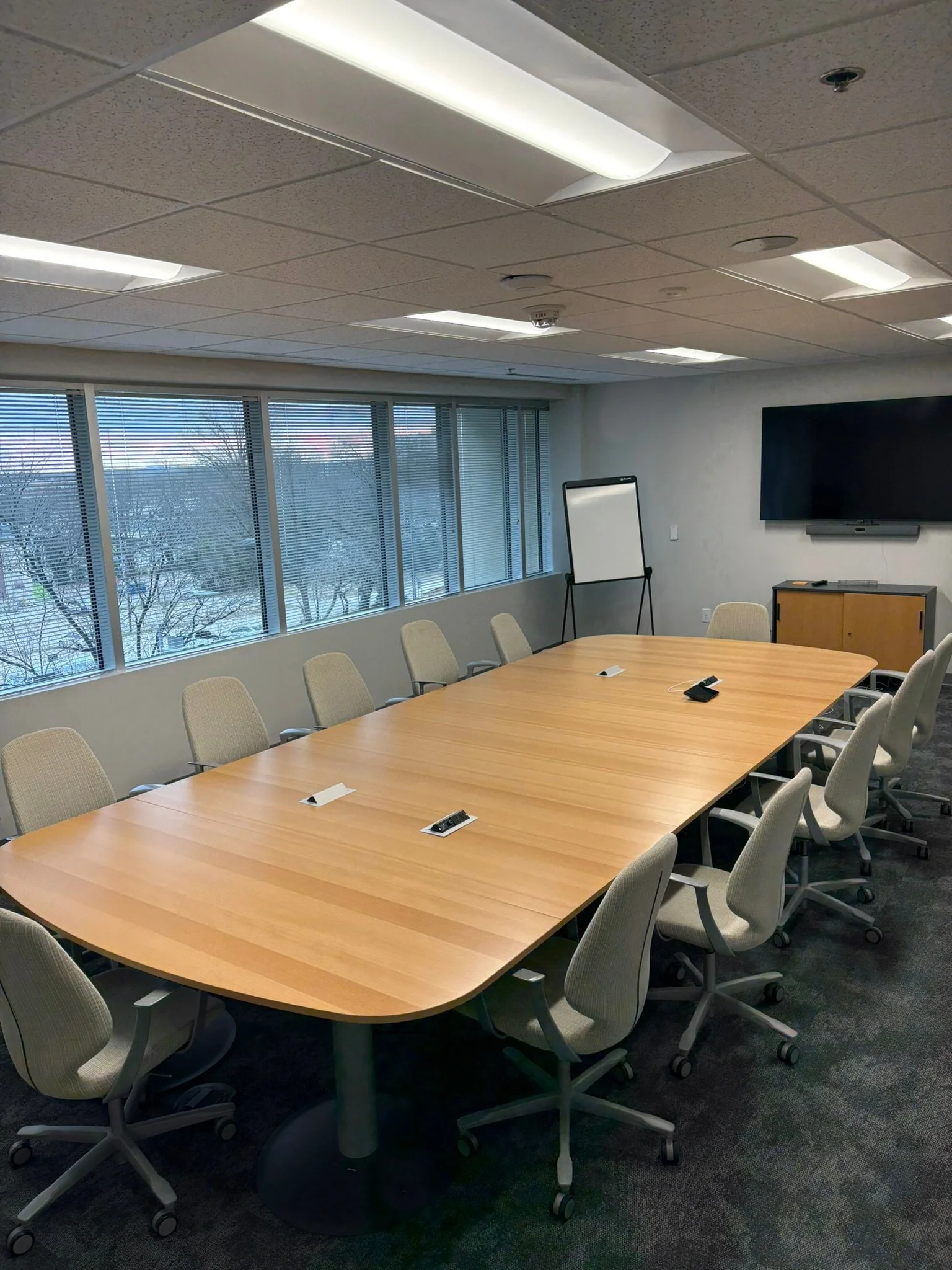 A conference room with a long wooden table surrounded by beige office chairs, a large window with blinds, a whiteboard, a wall-mounted television, and ceiling lights.