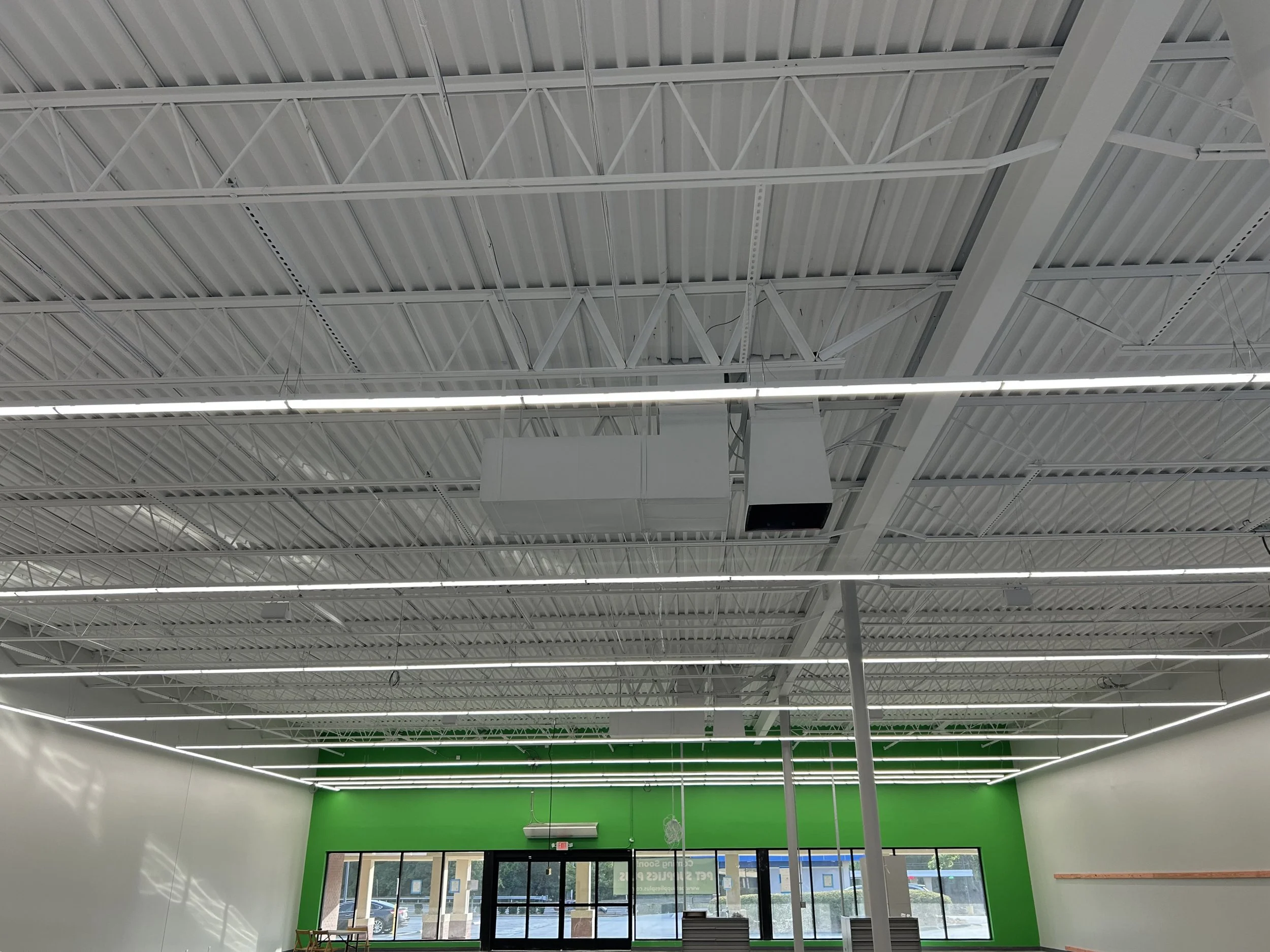 Interior view of a store with a high metal ceiling, fluorescent lighting, and a green accent wall near the entrance. Large glass doors and windows at the front allow natural light inside.