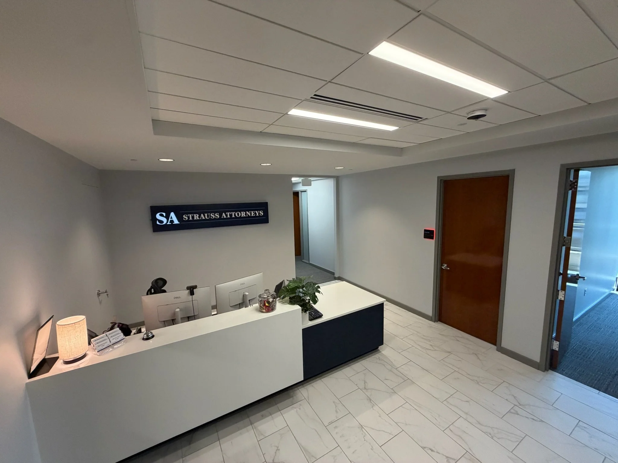Office reception area with a white curved reception desk displaying two computer monitors, a lamp, and office supplies. There are two chairs behind the desk, a plant in a glass container, and a sign that reads 'SA Strauss Attorneys'. The ceiling has recessed lighting and white tiles, and there is a wooden door on the right side.