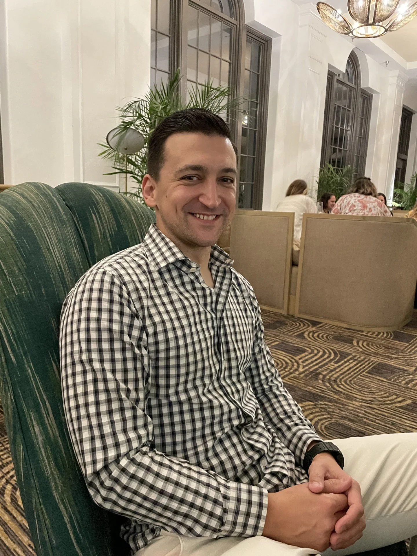 A man smiling, wearing a checkered shirt, seated in a restaurant with a green upholstered chair, large windows, indoor plants, and other diners in the background.