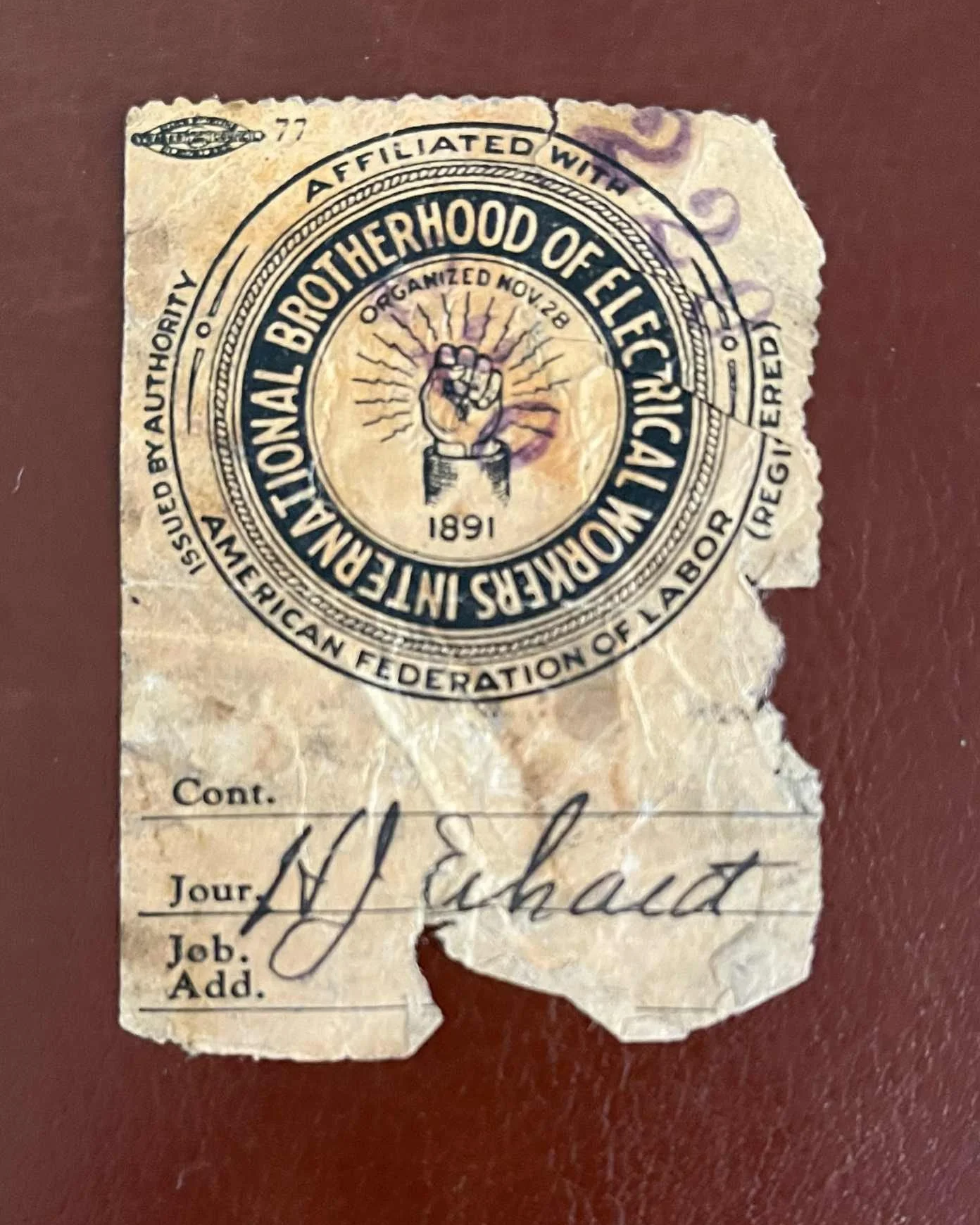 A torn and aged receipt with the emblem of the Brotherhood of Electric Workers, featuring a raised fist and the words 'Organized Nov 28' and '1891,' on a dark red surface.