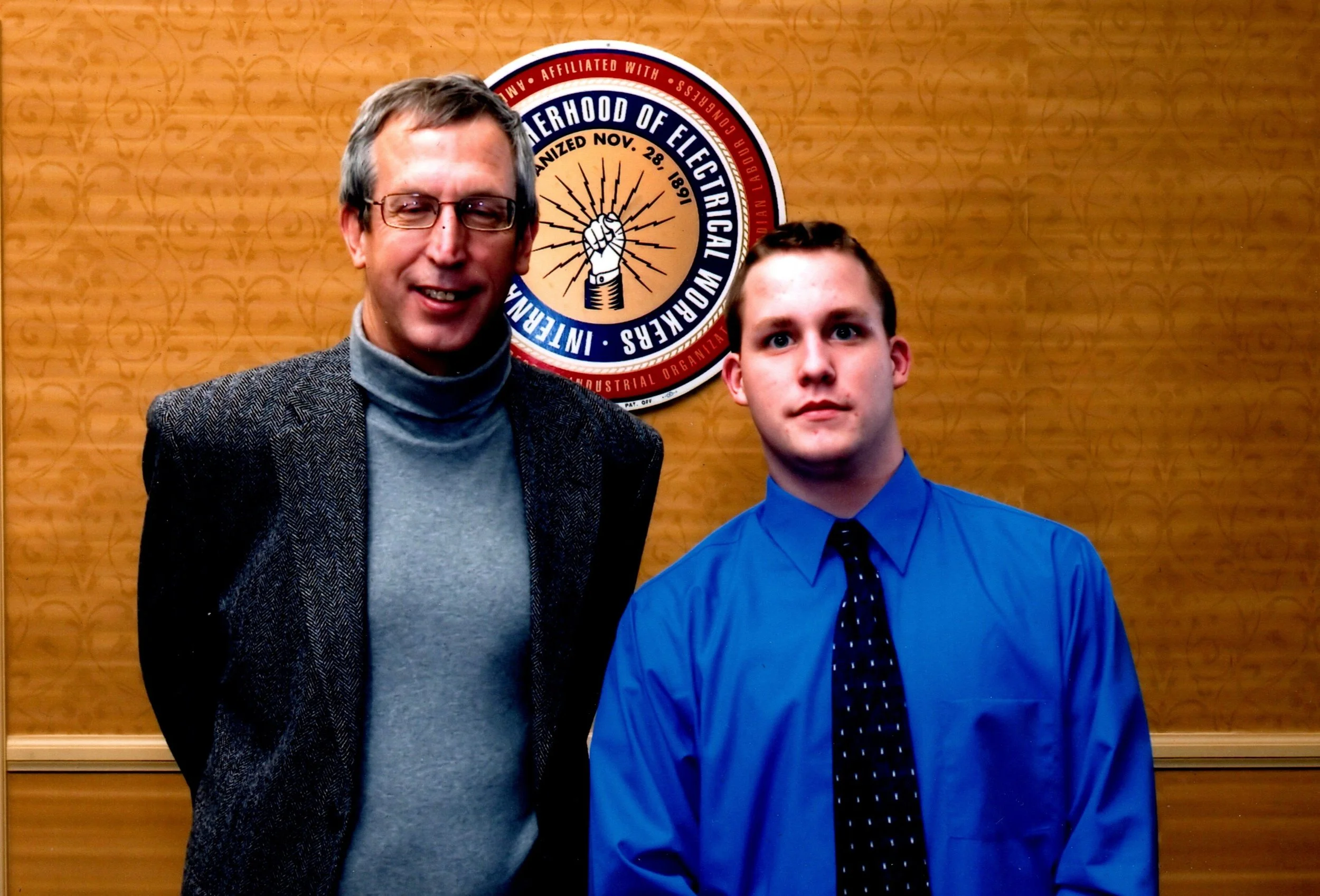 Two males, one older with glasses and gray hair, in a gray turtleneck and blazer, and a younger male with short brown hair, in a blue shirt and black tie, standing in front of a wooden wall with a circular emblem that reads "Brotherhood Of Electrical Workers".