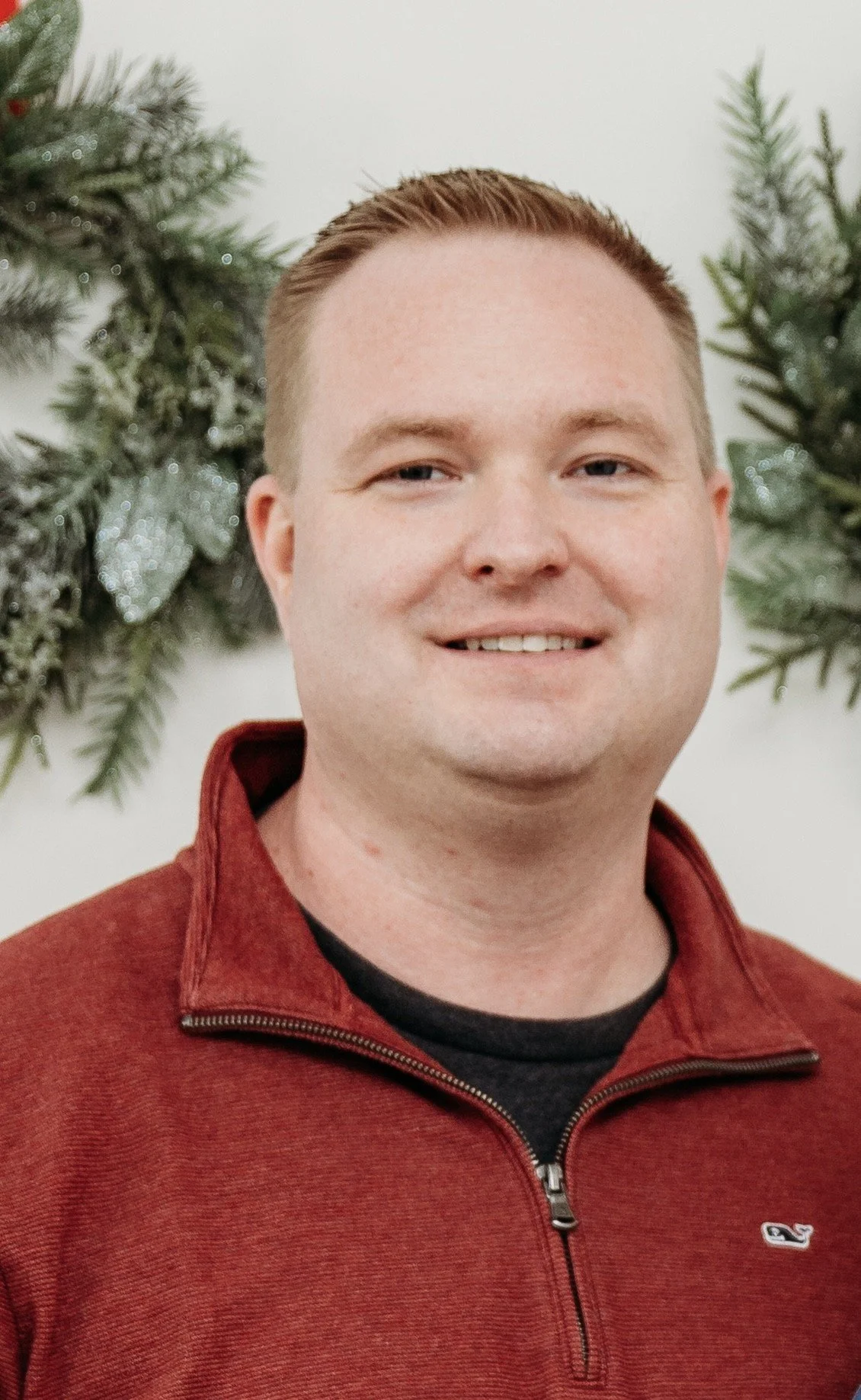 A smiling man with short red hair wearing a maroon zip-up jacket and a black shirt, standing in front of Christmas greenery on a wall.
