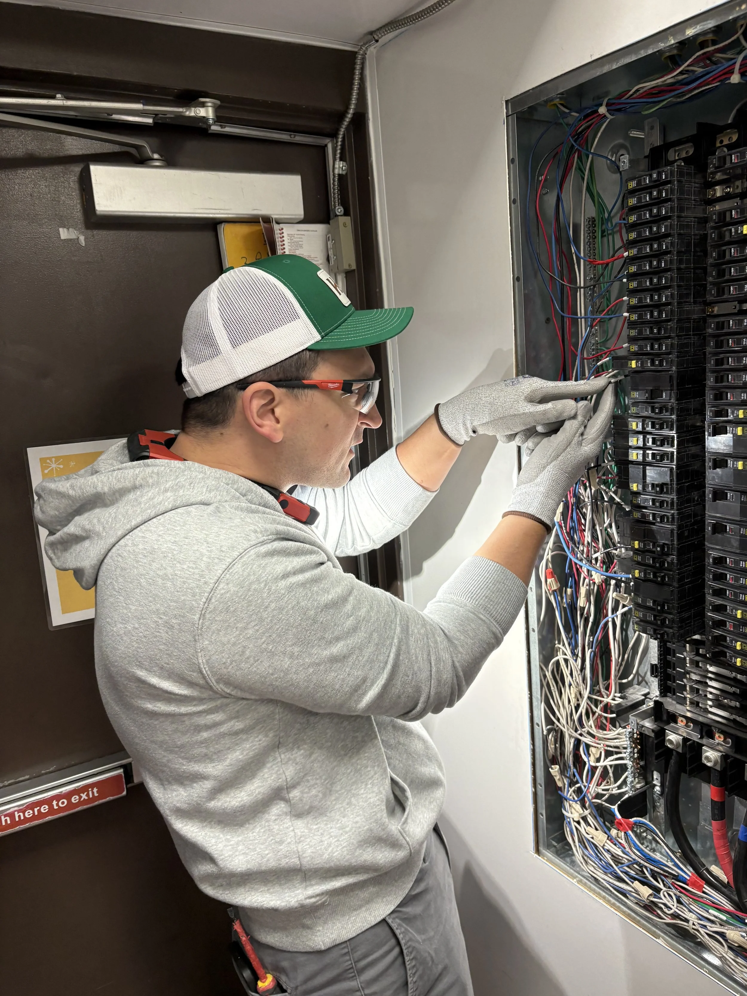 A technician wearing safety glasses, a baseball cap, and gloves working on an electrical panel with multiple wires.