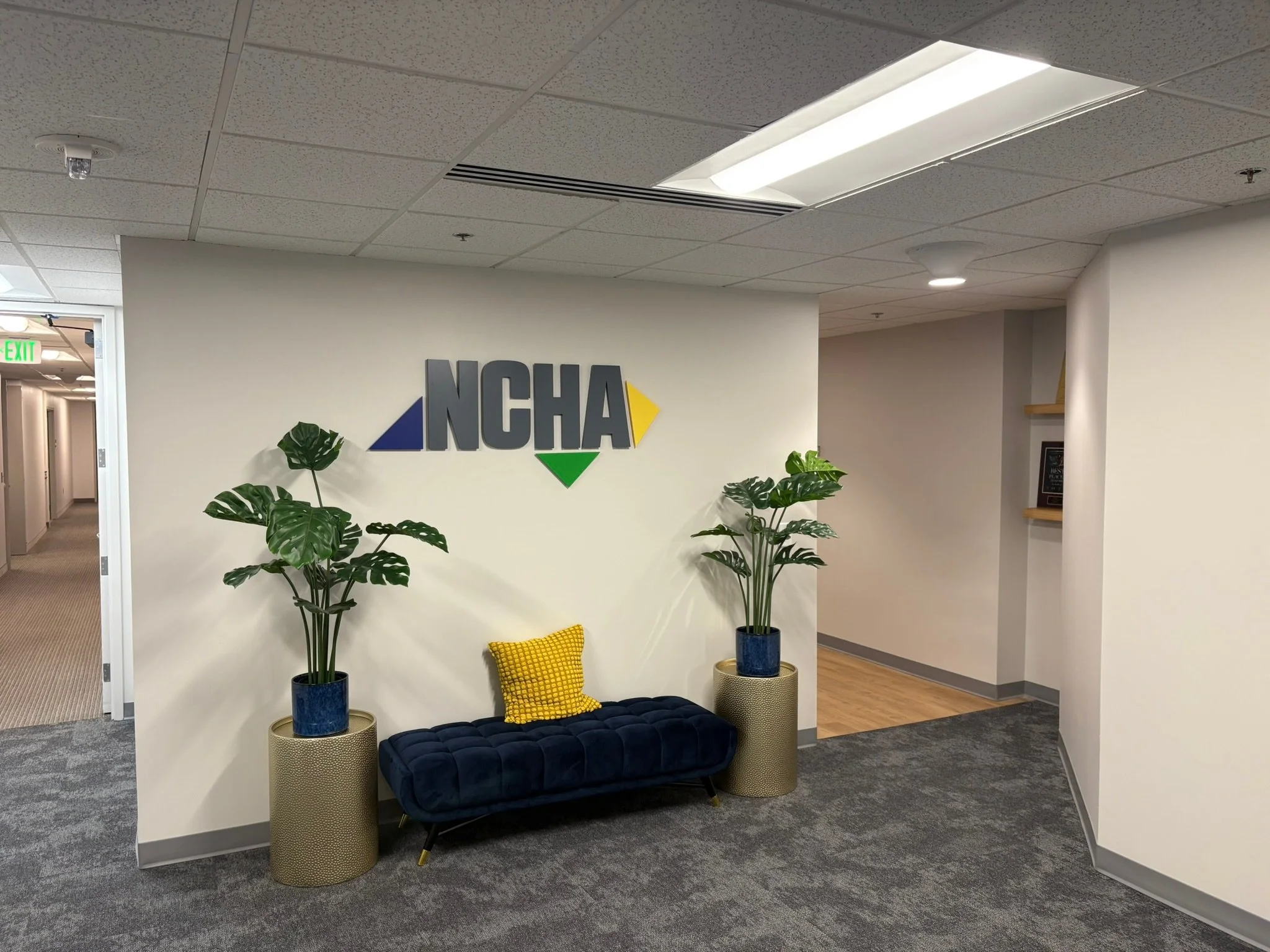 NCHA's office lobby with plants, a navy blue velvet bench, a yellow patterned pillow, and a sign with the letters NCHA on the wall.