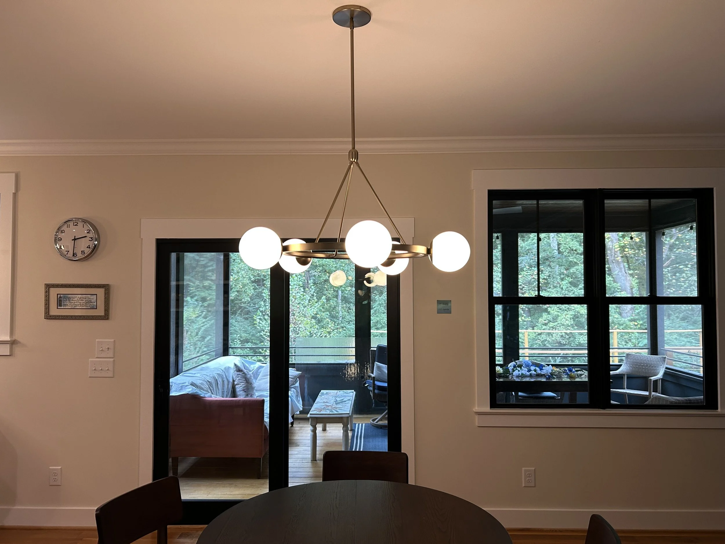 Interior view of a dining room with a round black table and chairs, a modern chandelier with white globe lights hanging from a ceiling, sliding glass doors opening to a patio with trees outside, a wall clock, and framed art on the wall.