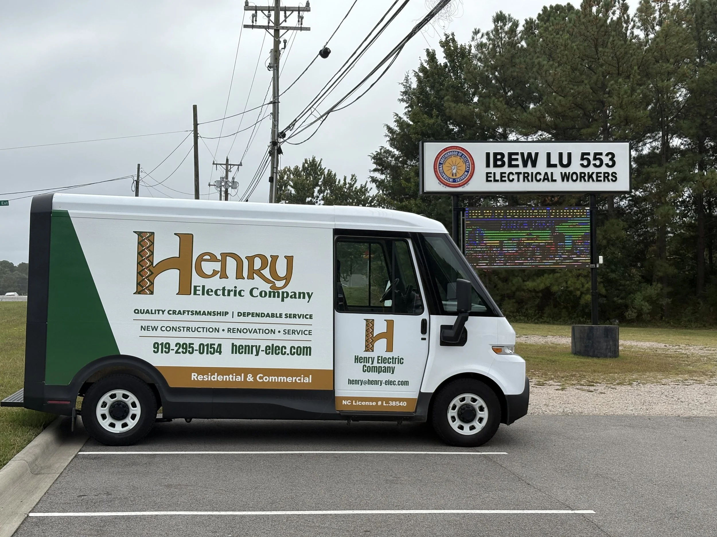 Electric company service van parked in a lot with a sign behind it that reads "IBEW LU 553 Electrical Workers" and a logo, with utility poles and trees in the background.