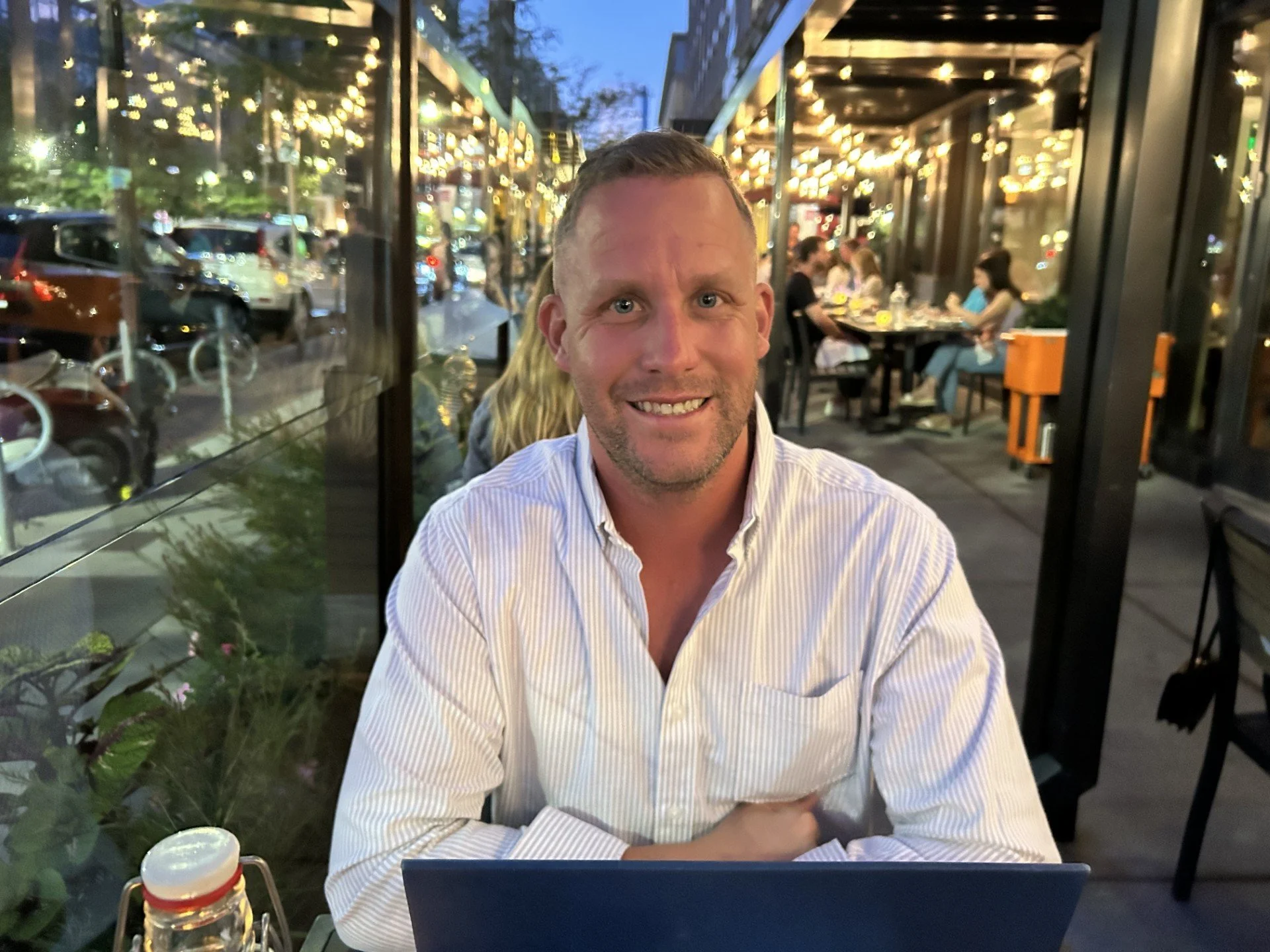 A man in a white striped shirt sitting outdoors during evening, smiling at the camera with a laptop in front of him. There are people dining inside a restaurant with string lights, parked cars, and bicycles visible through the glass window.