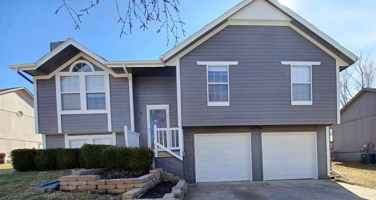 A two-story house with gray siding, white trim, and a split garage with two doors. The front entrance has a small staircase and a white door, and there are three large windows on the upper level and two on the lower. There are bushes and a small stone retaining wall in the front yard.