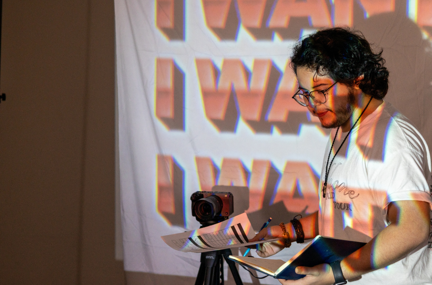 A young man with curly dark hair, glasses, and a beard, standing in front of a white background with a colorful, abstract pattern projected on it. He is holding a notebook and a pen, standing next to a camera on a tripod, with some papers in front of him.