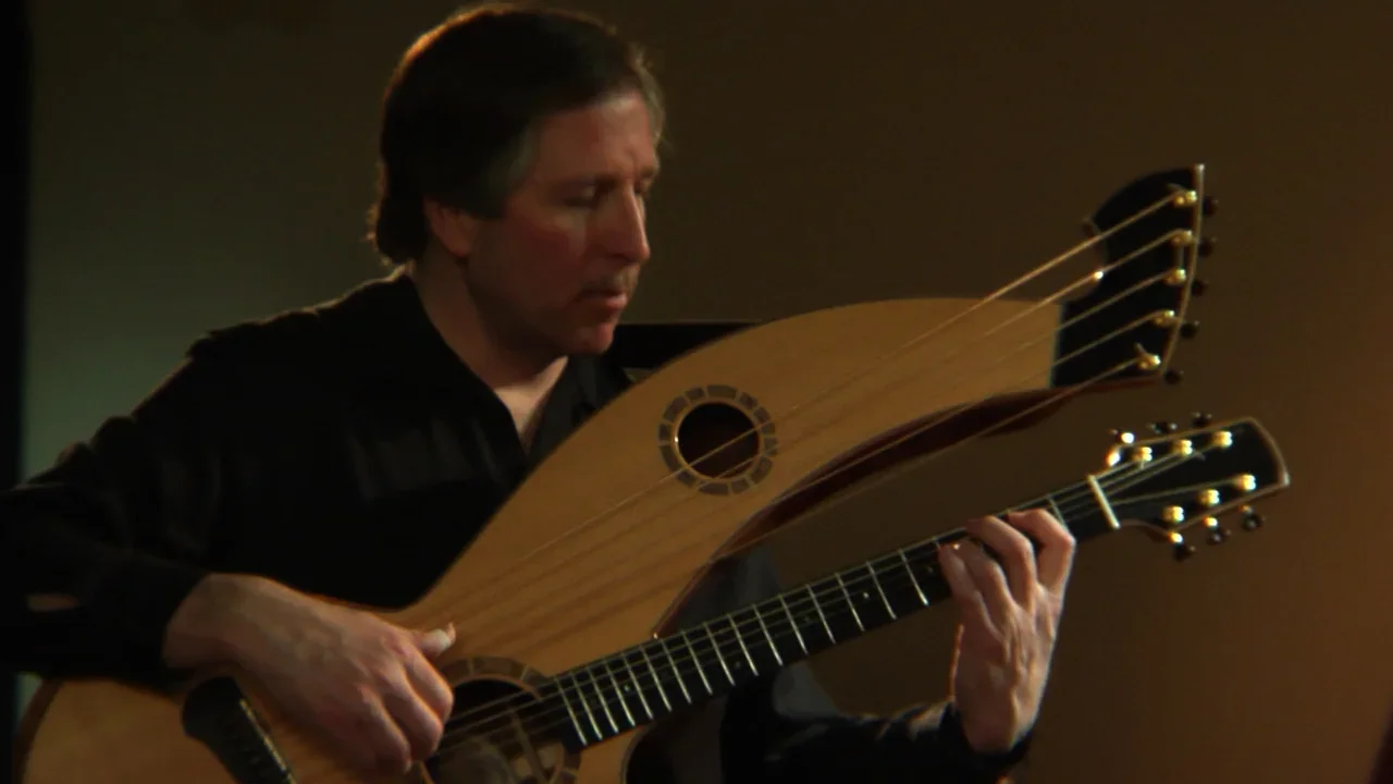 A man playing a hybrid guitar that combines a classical guitar and a harp, seated and focused on his playing.