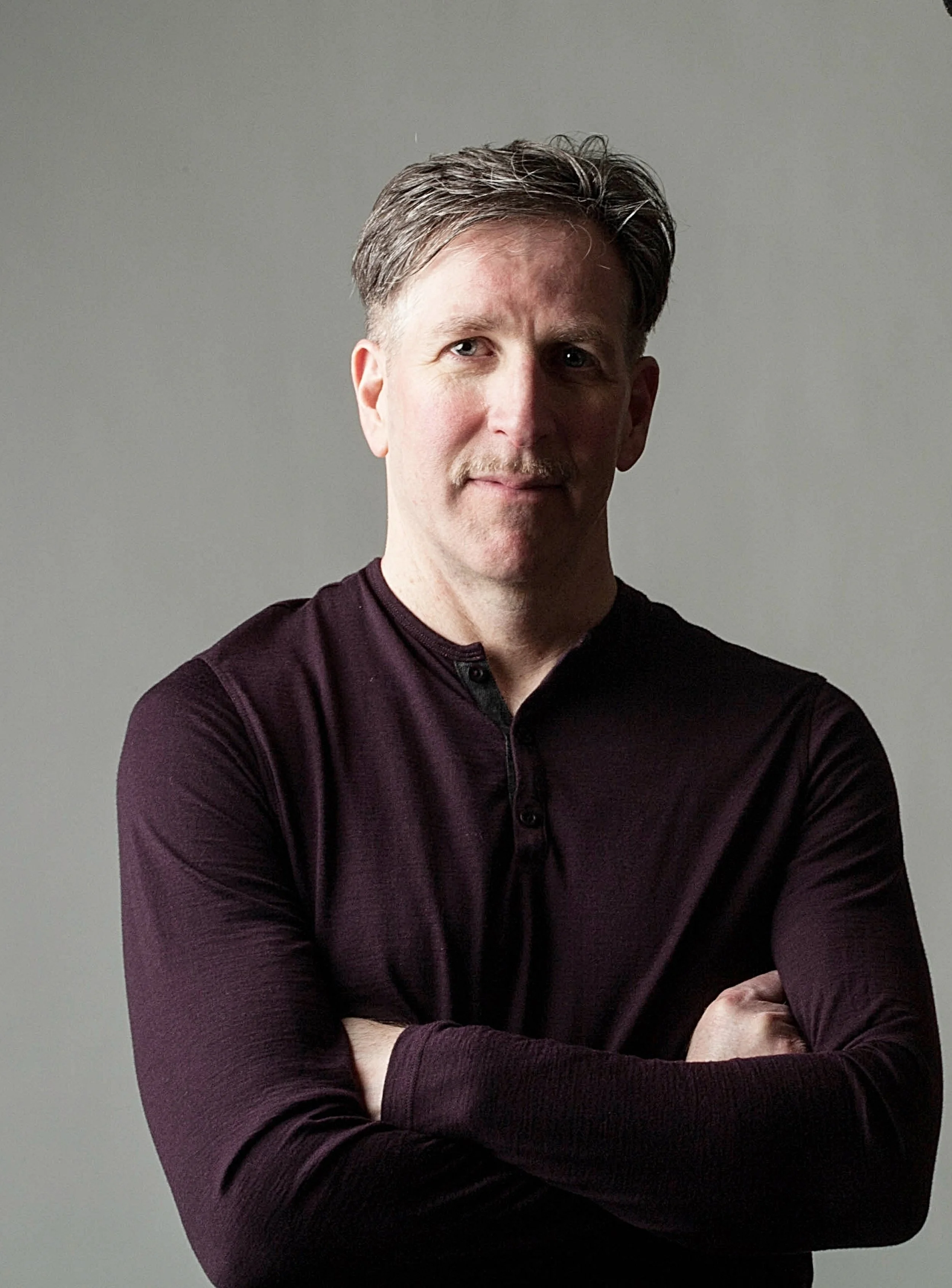 Mark Vickness. Portrait of a man with crossed arms, light gray background, wearing a dark long-sleeve shirt.