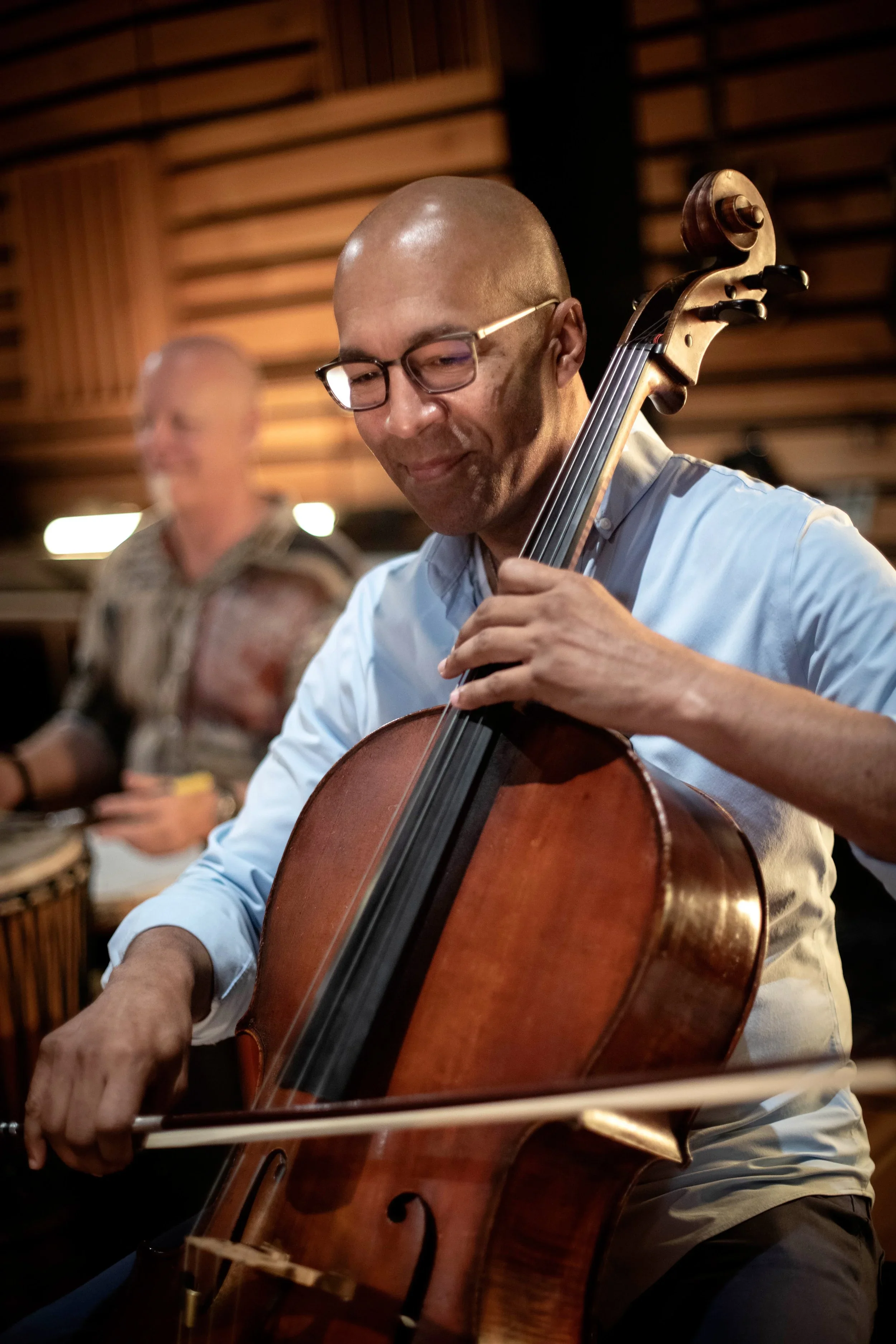Joseph Hebert. A man playing an upright bass in a music studio with wooden walls, with another musician in the background.