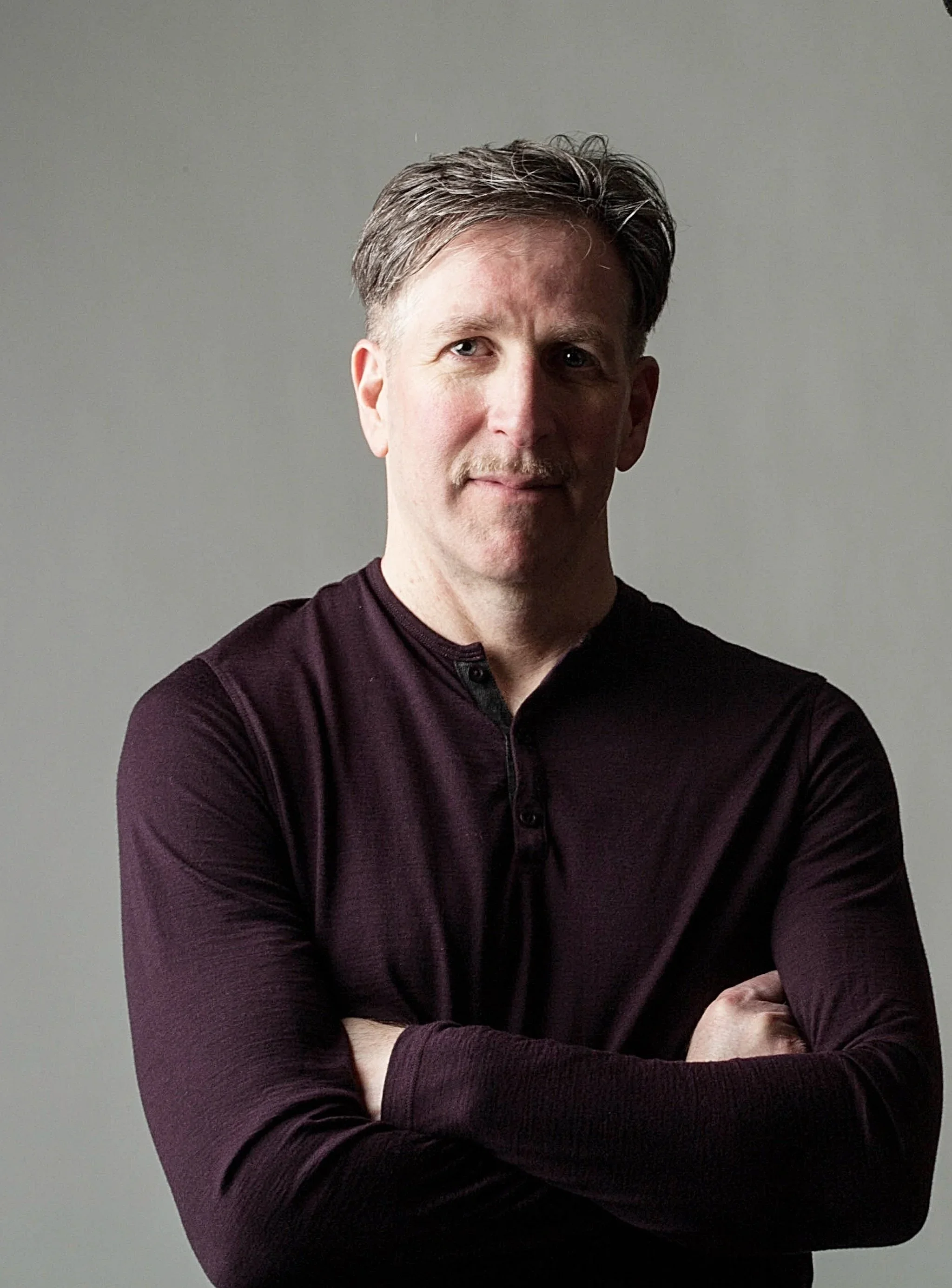 A middle-aged man with short light brown hair and a mustache, crossing his arms and looking at the camera, wearing a dark long-sleeve shirt against a plain gray background.