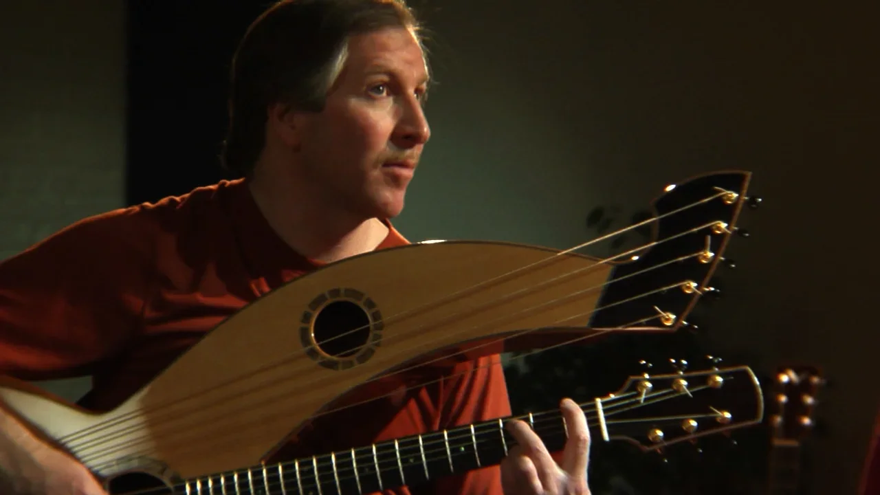 Mark Vickness playing a multi-neck acoustic guitar in a dimly lit room.
