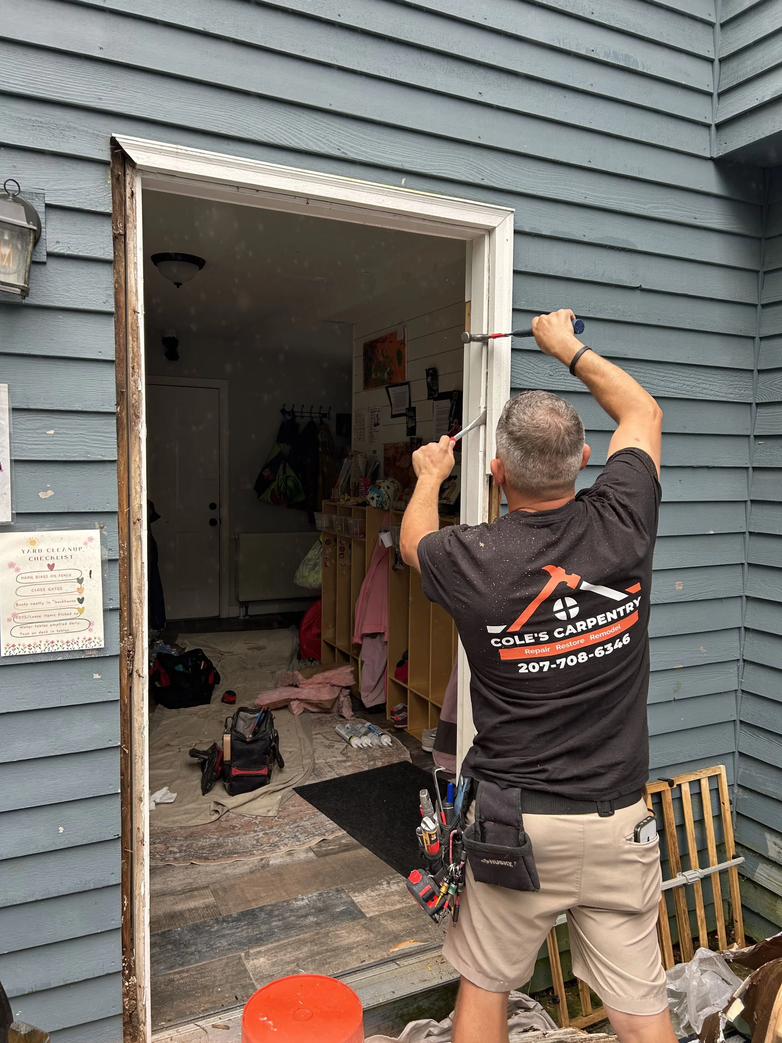 A man in a black shirt and beige shorts is working on installing or repairing a window frame on a light blue house. He has tools attached to his belt and is using a screwdriver. Inside the house, clutter and tools are visible, including a black bag, bottles, and some clothing or fabric on the floor.