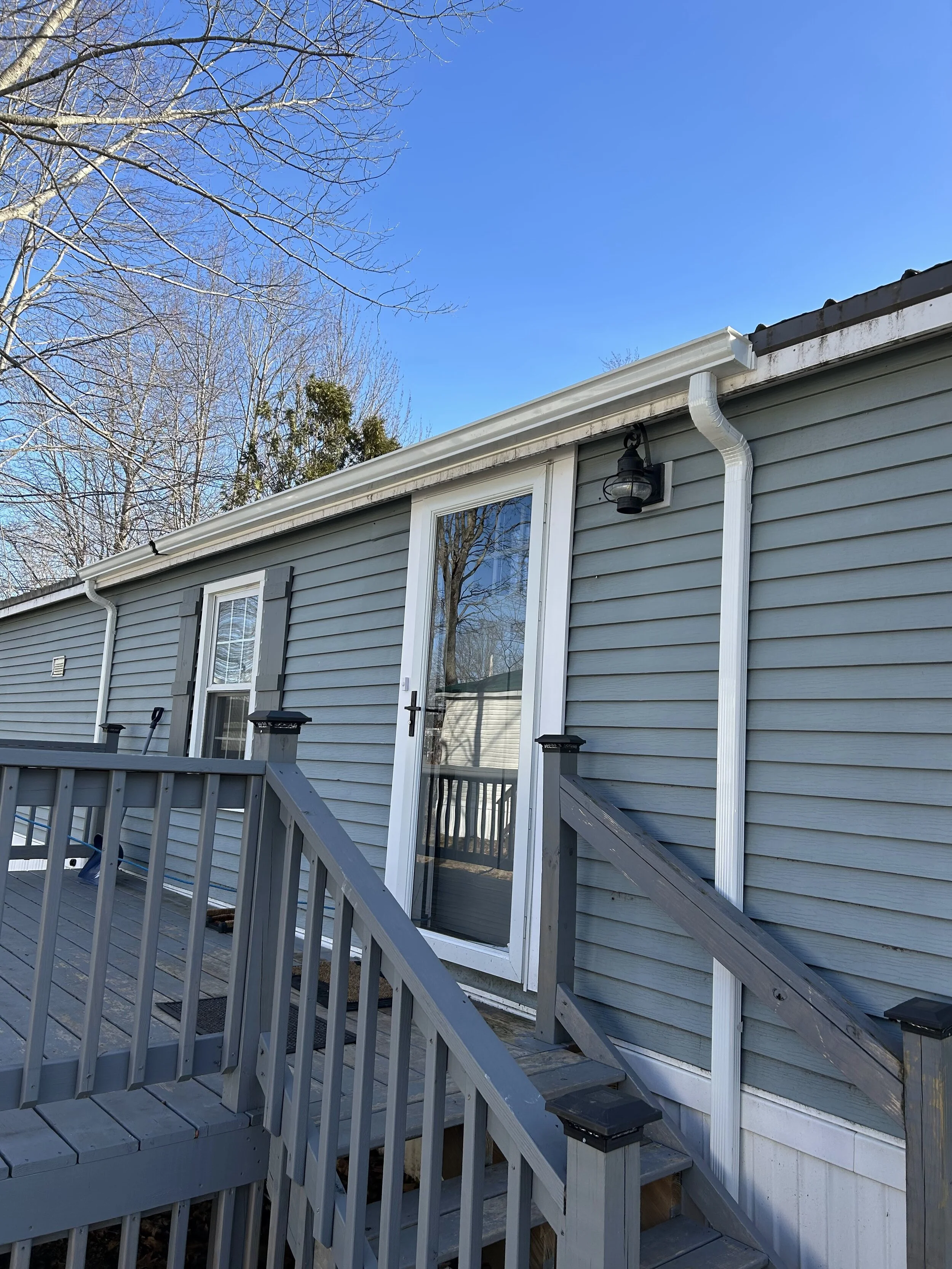 Back porch of a house with gray siding, a glass door, and stairs with a railing, set against a clear blue sky with bare trees in the background.
