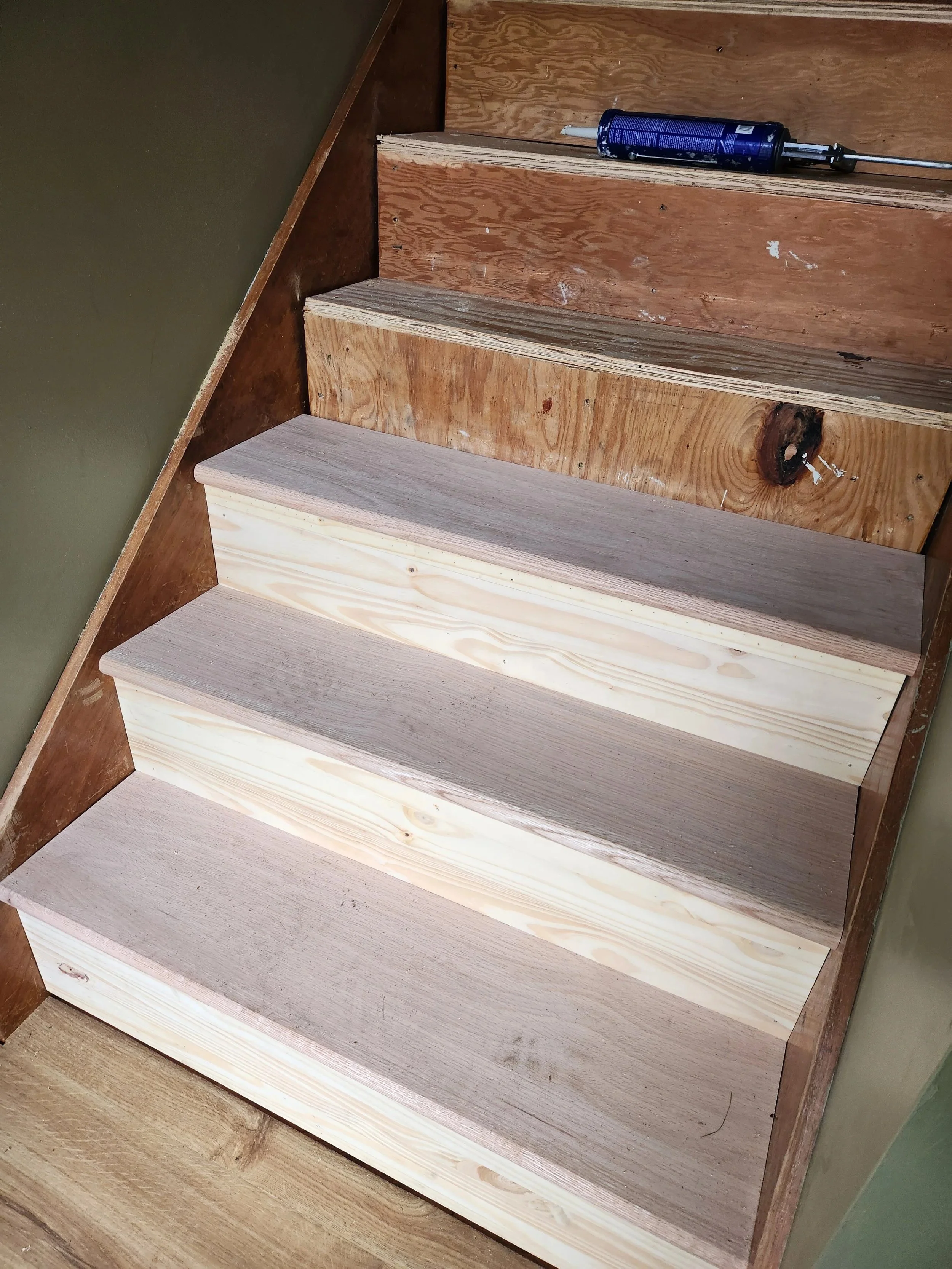Wooden staircase with four steps, some sanded and some still rough, with a bottle of construction adhesive and a caulking gun on the top step.