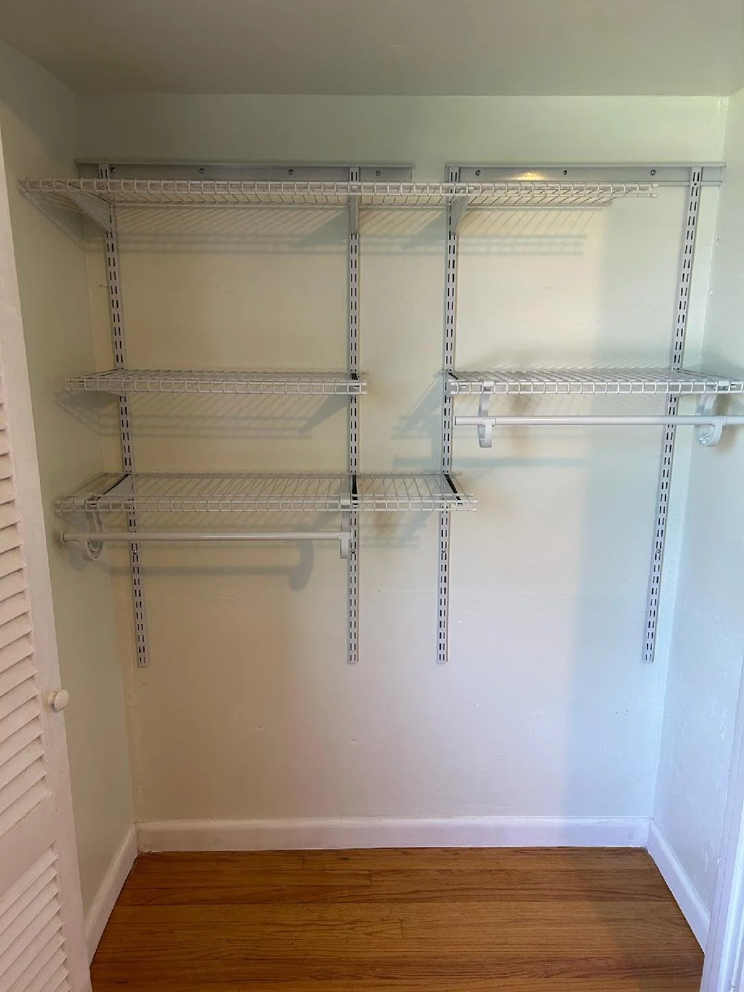 Empty white wire shelving unit with three shelves and a clothing rod, installed in a closet with light green walls and wooden floor.