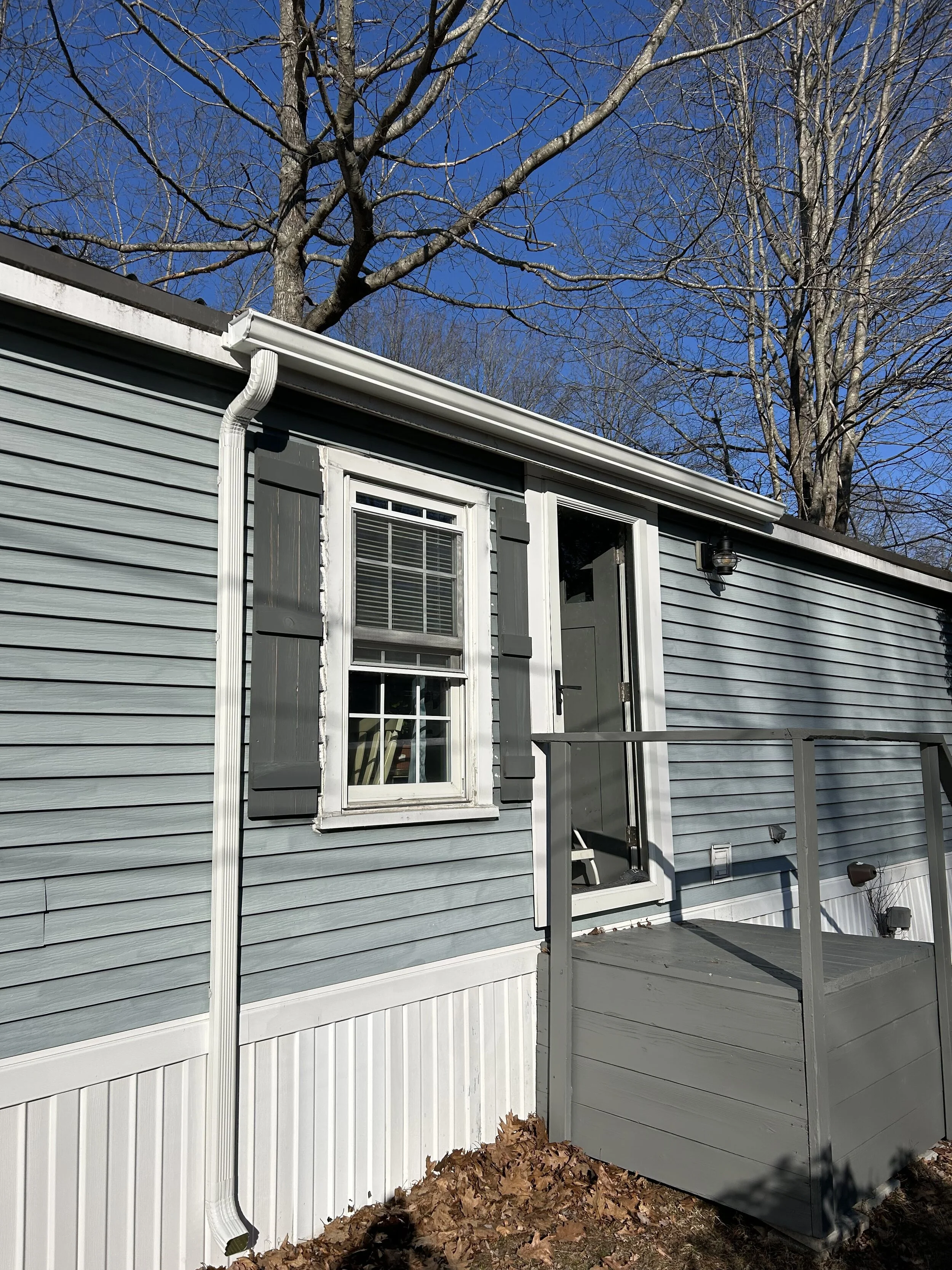 Exterior view of a house with blue siding, a window with black shutters, a balcony with a door, a white gutter, and leafless trees in the background under a clear blue sky.