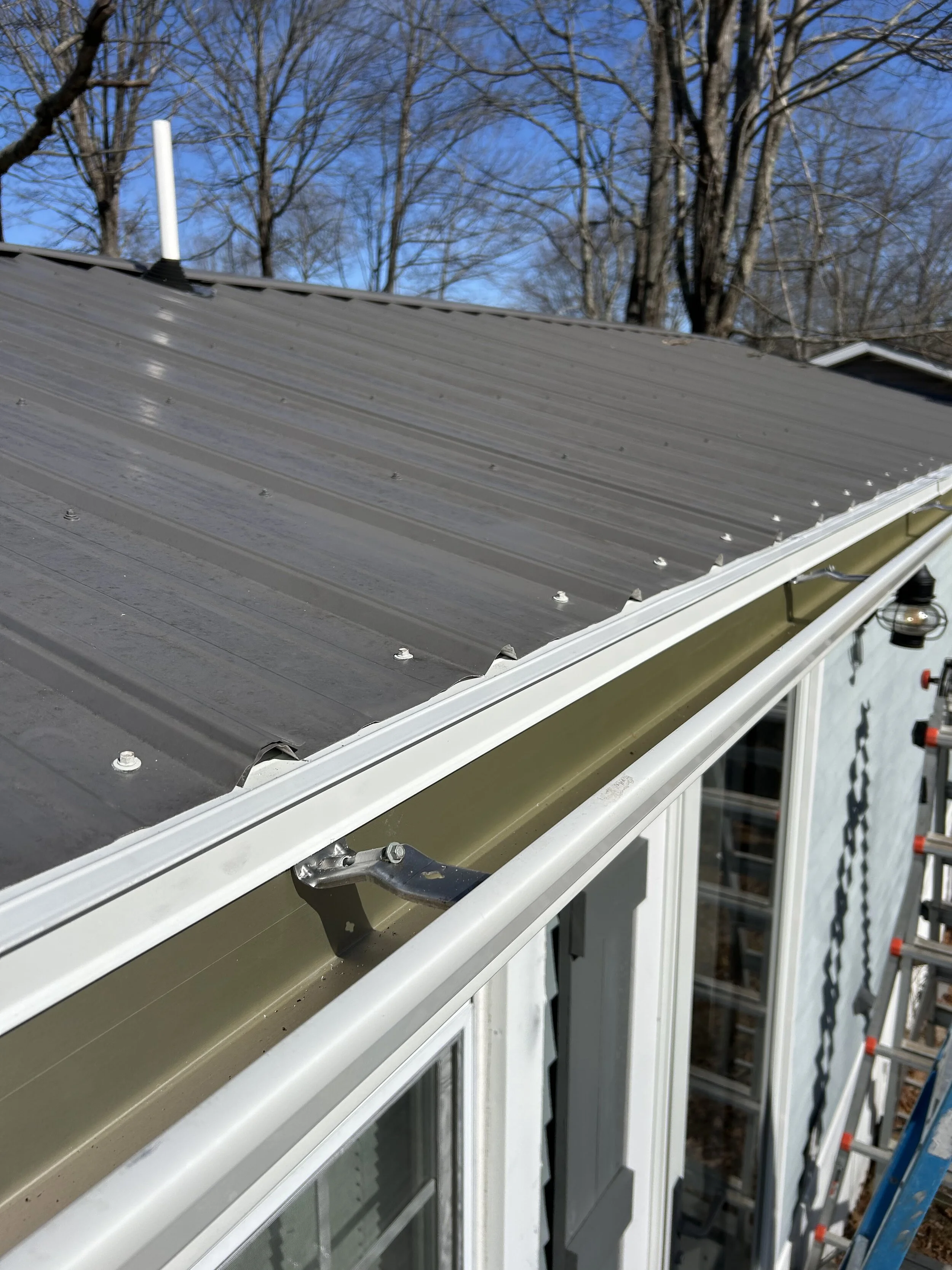 Close-up of a metal roof being installed on a house, with a ladder and trees in the background.