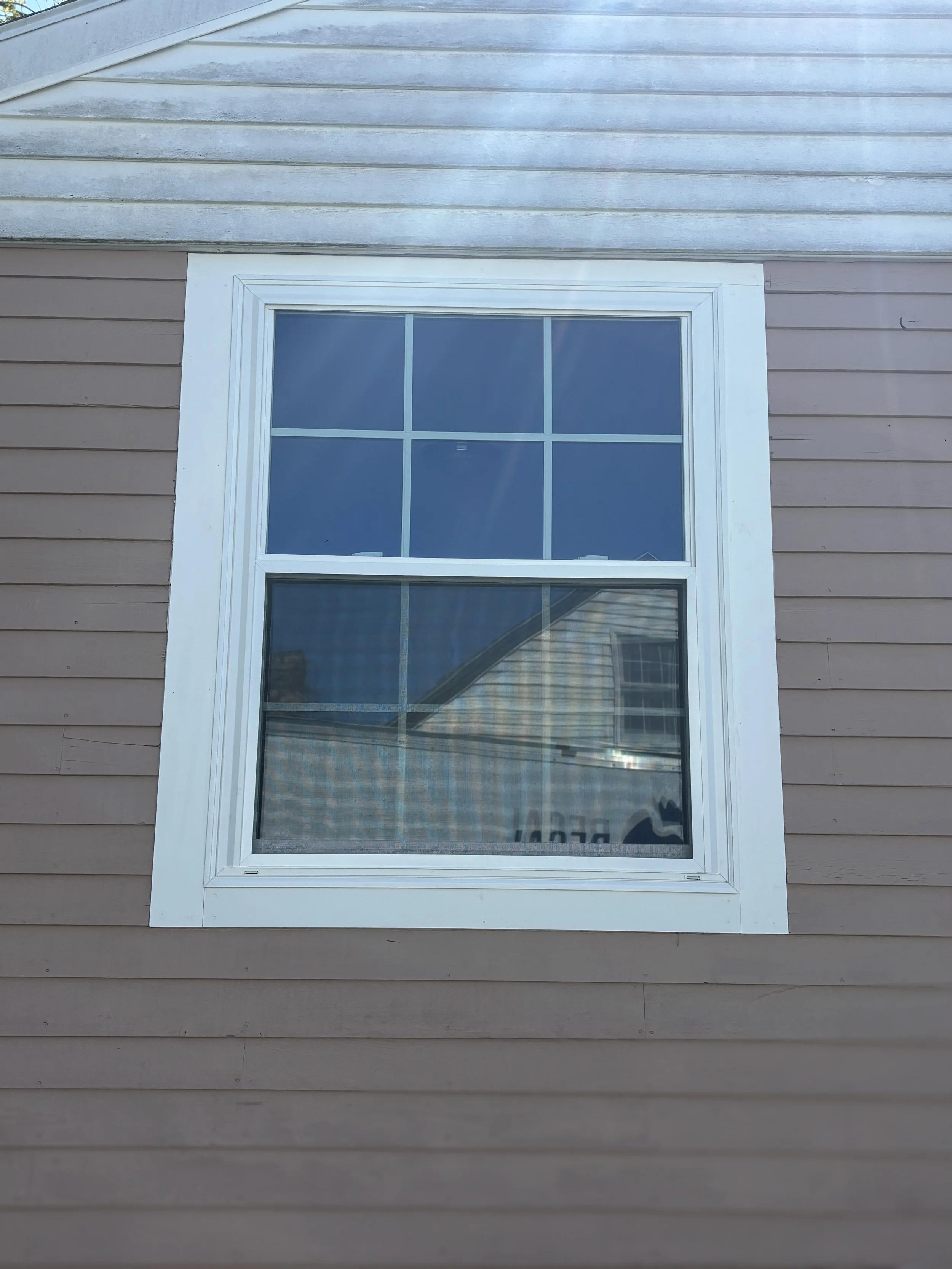 Close-up view of a double-hung window on the beige siding of a house, with the window divided into six panes and reflections of the sky and neighboring house visible.