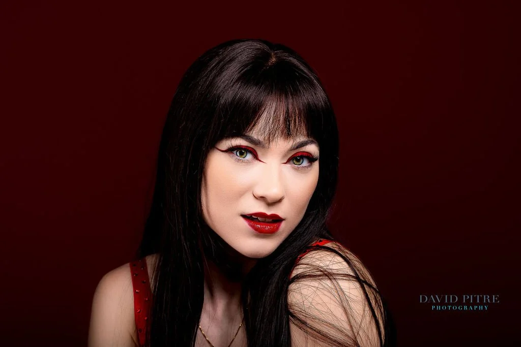 Close-up portrait of a woman with black hair, red lipstick, and makeup, against a dark red background.