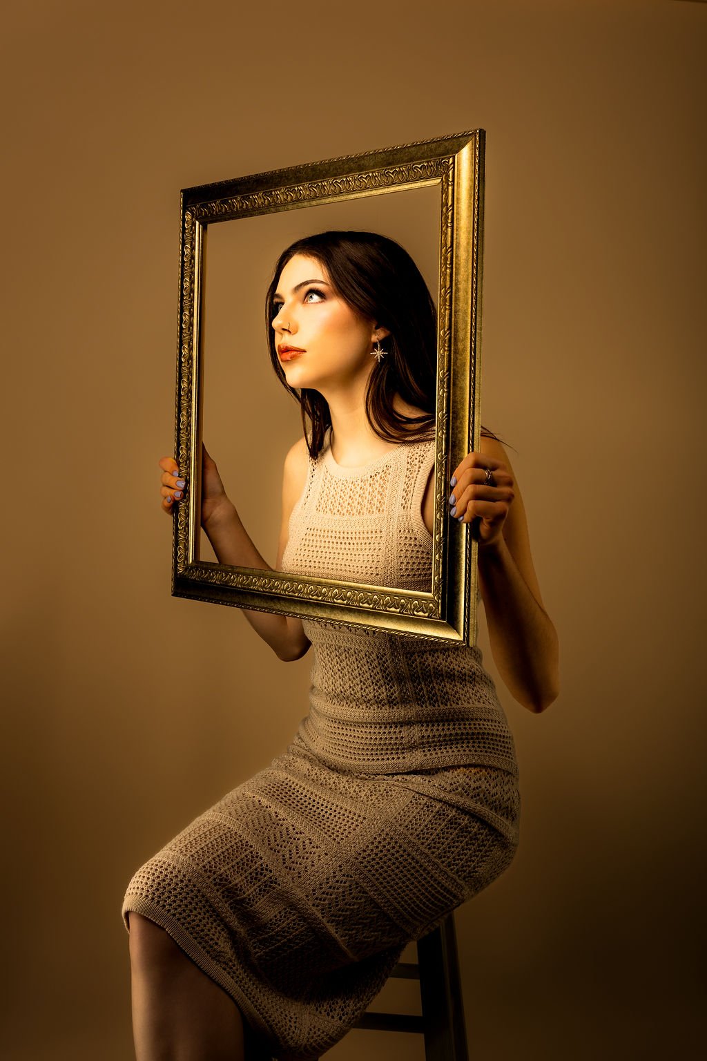 A woman with dark hair and earrings holding an ornate gold picture frame in front of a brown background.