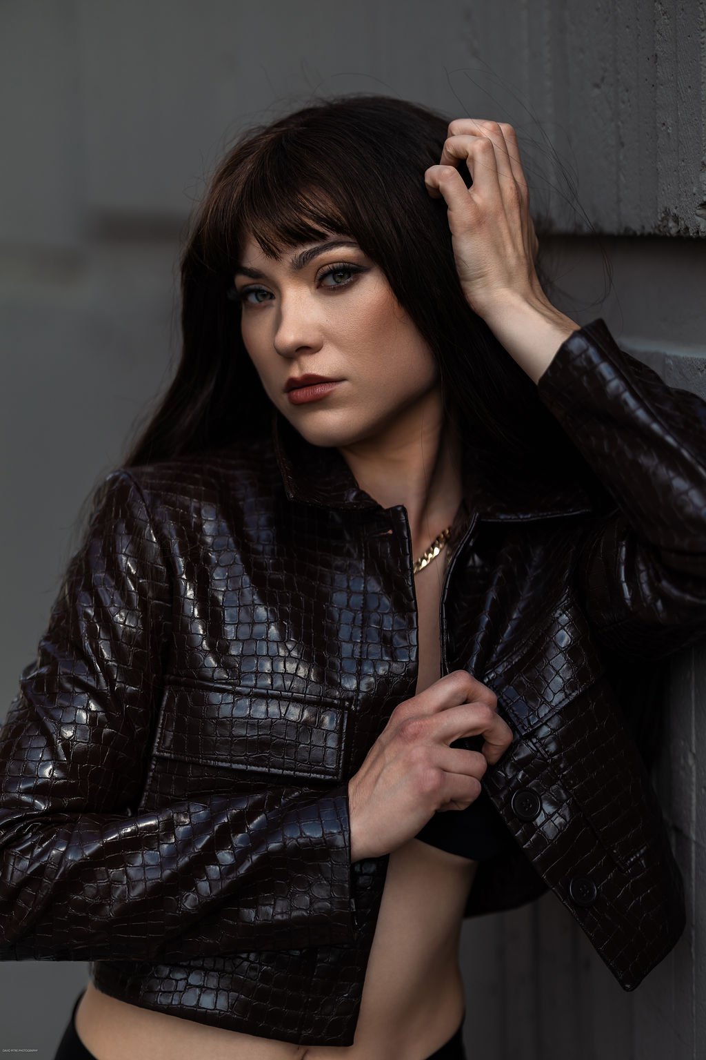 A woman with dark brown hair, wearing a black crocodile-pattern leather jacket, standing against a gray wall, looking into the camera.