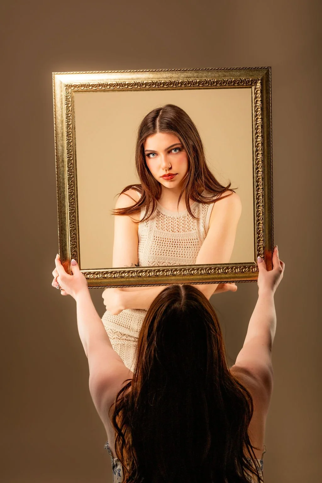 A woman holding a framed mirror, reflecting another woman with long brown hair, blue eyes, and a beige sleeveless knit top.