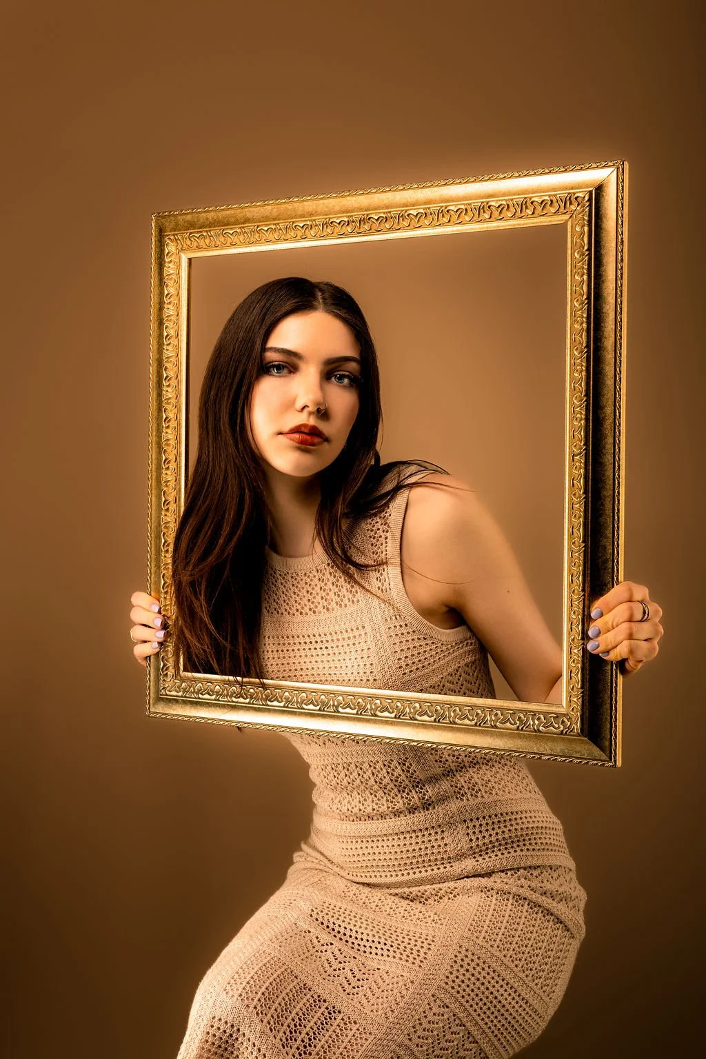 Young woman with long dark hair holding a large ornate gold picture frame, looking at the camera, with a plain brown background.