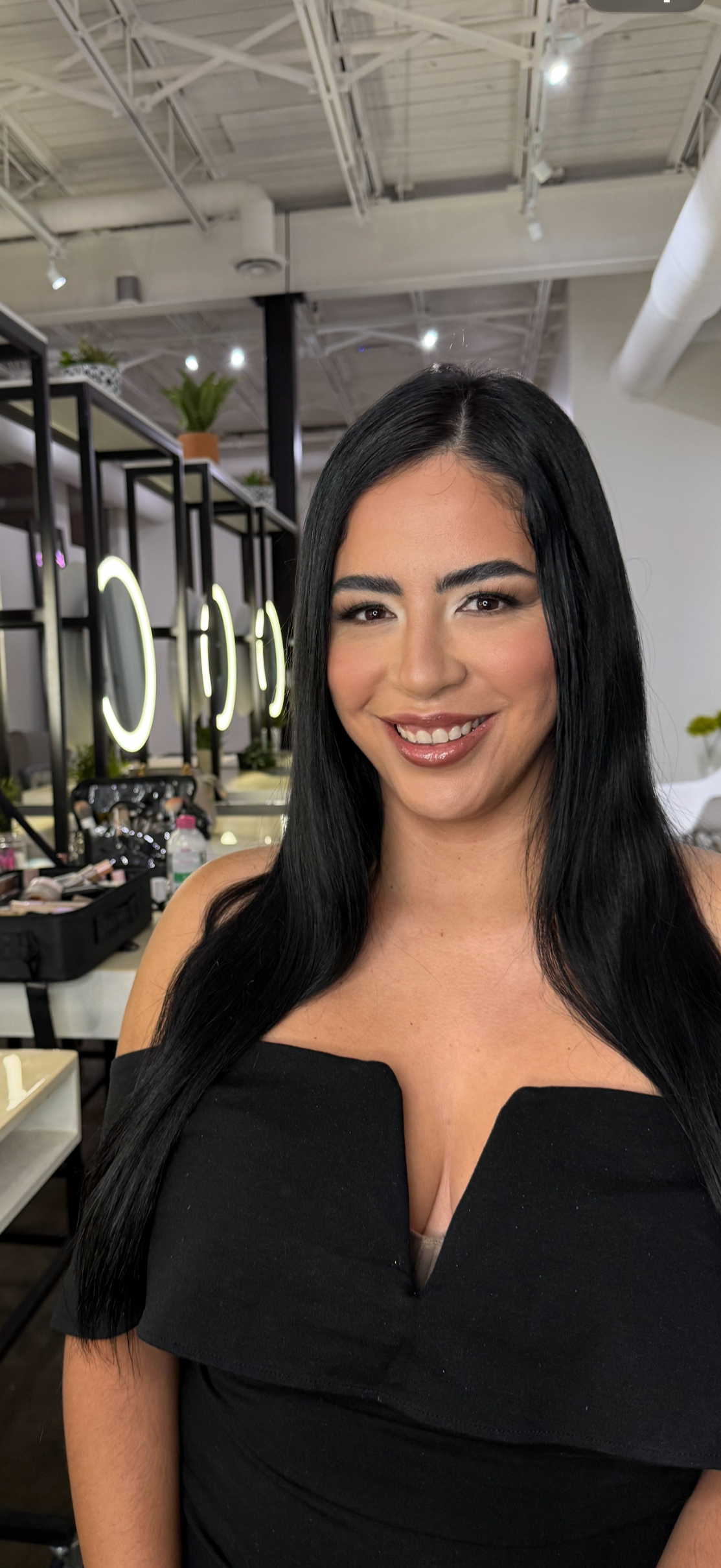 A woman with long black hair and a black dress smiling in a well-lit hair salon with makeup mirrors and makeup products in the background.