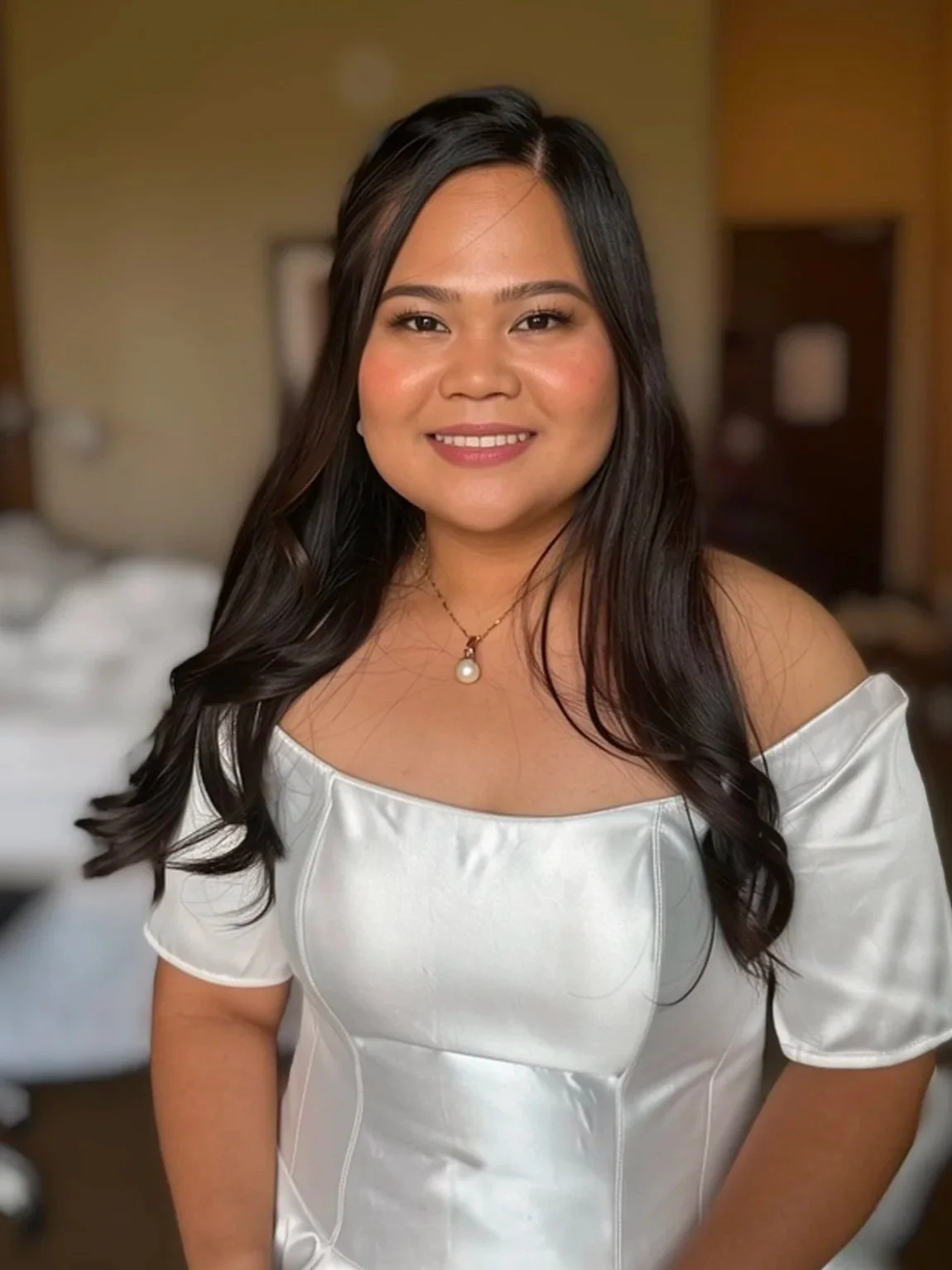 A woman with long wavy dark hair wearing a white off-the-shoulder satin dress and a necklace with a pearl pendant, smiling in an indoor setting.