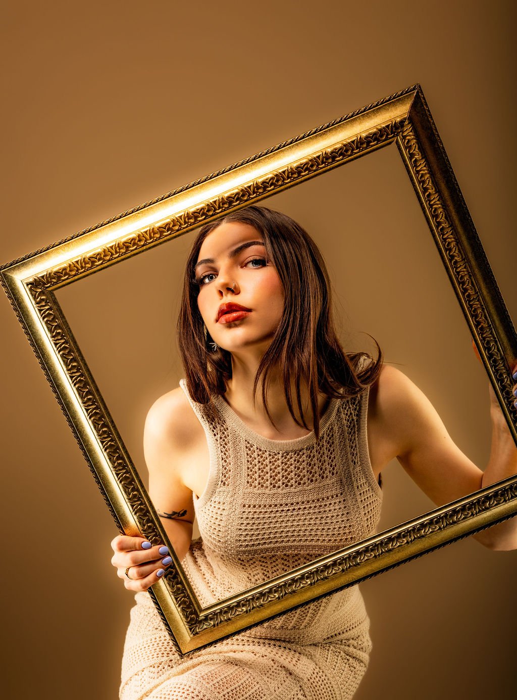 A young woman with brown hair and makeup holding a gold ornate picture frame in front of a brown background.