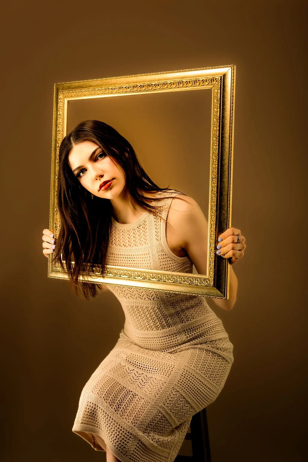 A young woman with dark hair and makeup, wearing a beige crochet dress, holding a gold picture frame with her head and shoulders inside, against a plain brown background.