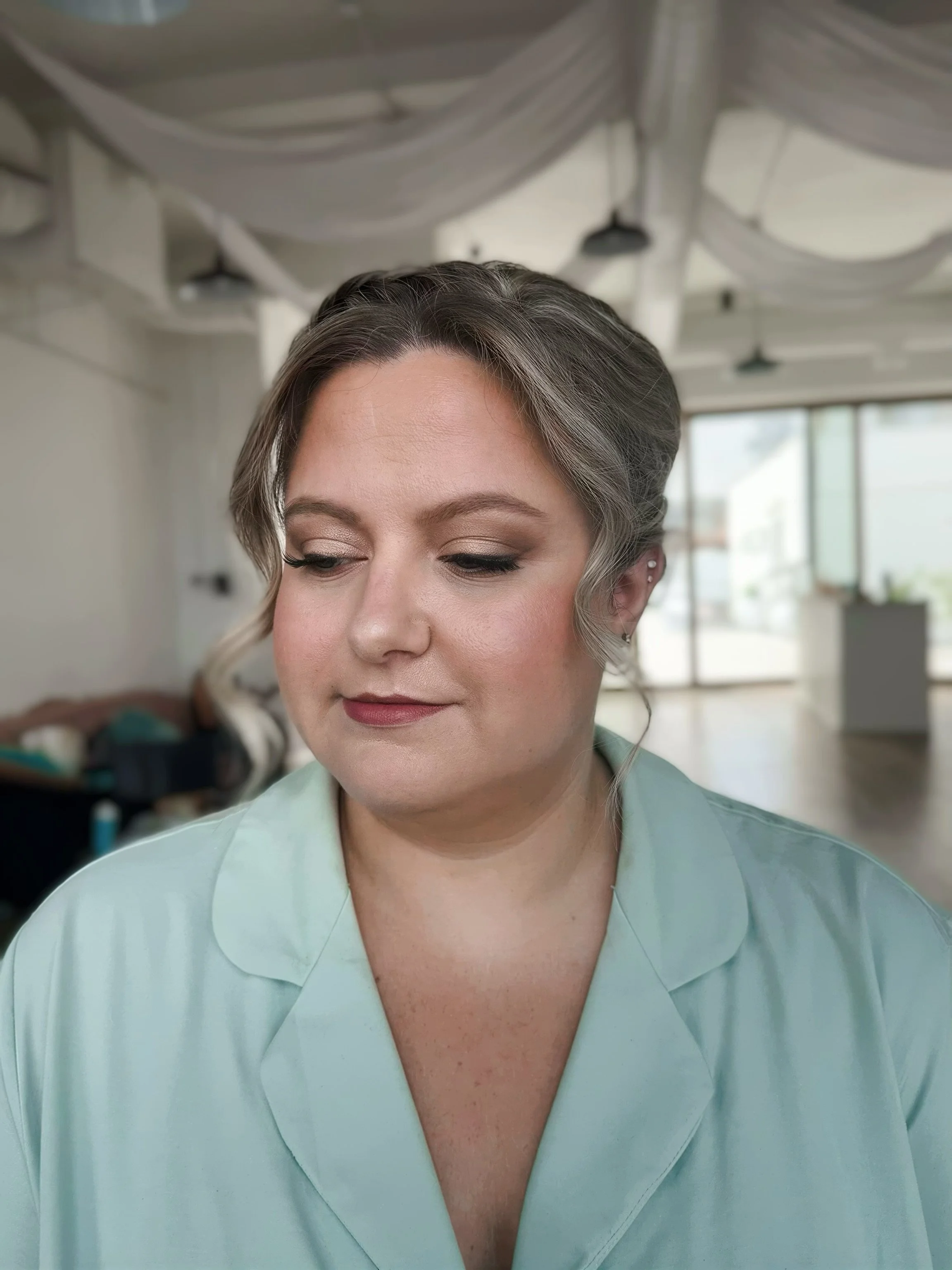 Close-up of a woman with styled hair and makeup, wearing a light green shirt, in a room with bright natural light and large windows.