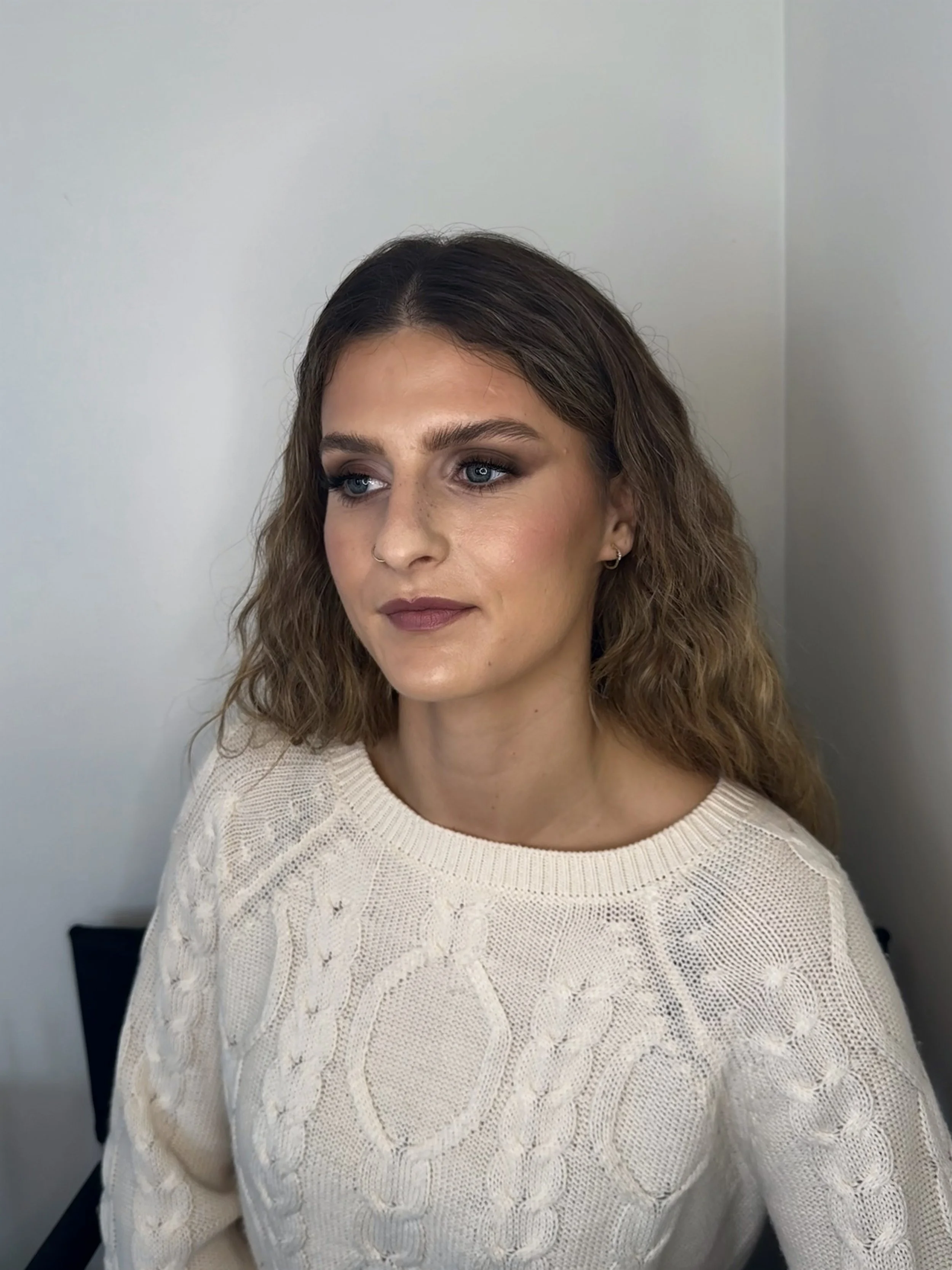 A woman with wavy brown hair and blue eyes, wearing a white knitted sweater with a textured pattern, sitting in front of a plain light-colored wall.
