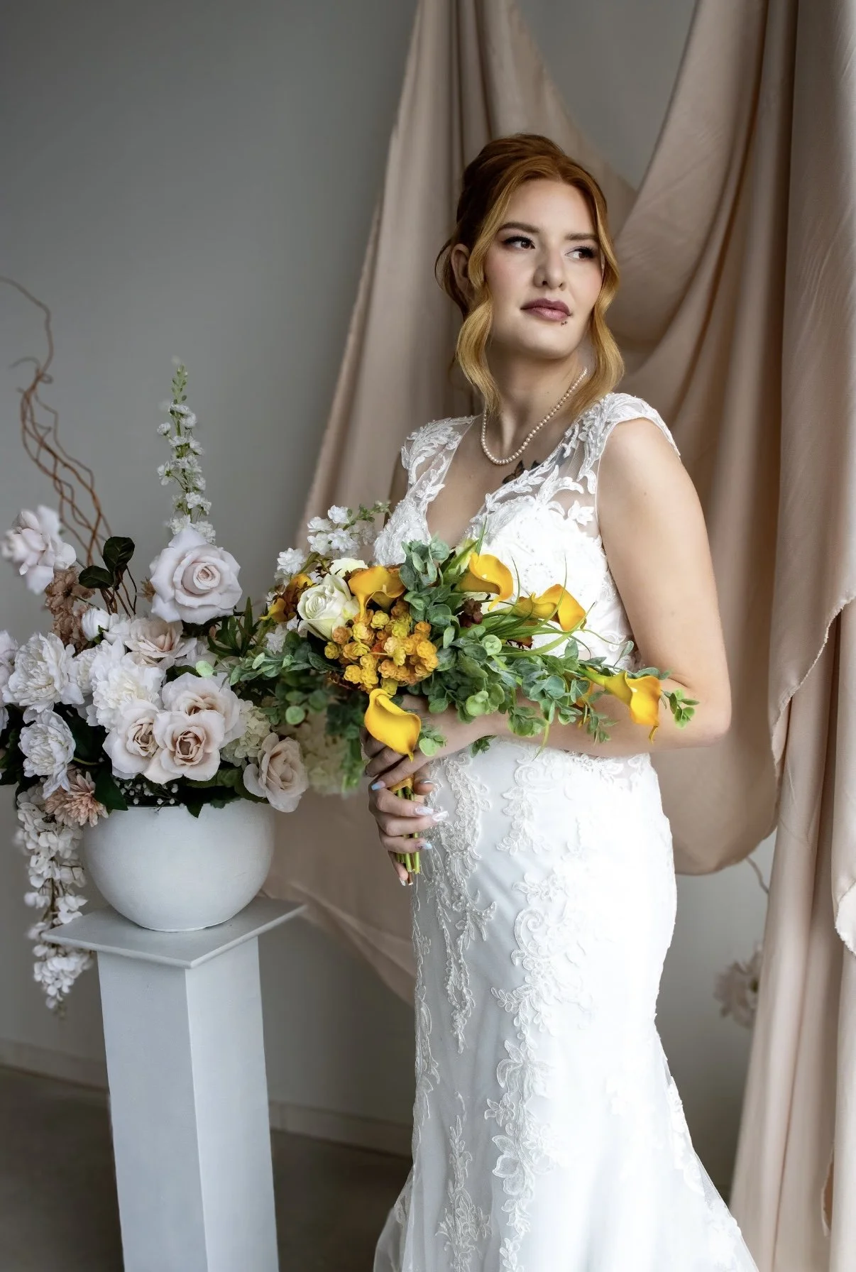 A bride with red hair in a white lace wedding gown holding a bouquet of yellow and white flowers, standing near a beige curtain and a white vase with pink and white flowers on a small white table.