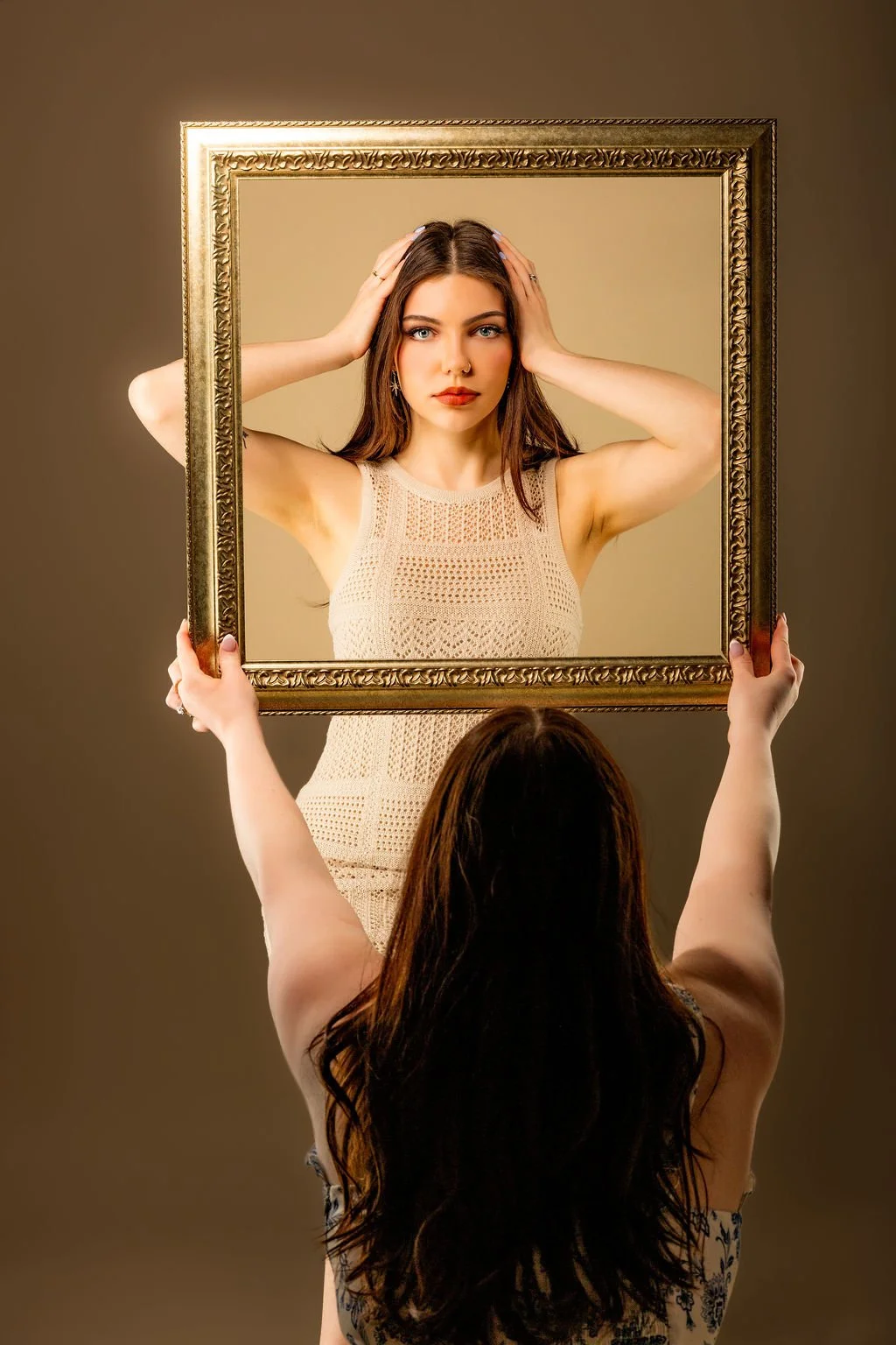 A woman looking into a mirror held up by another person, with her reflection showing a confident expression and hands on her head.