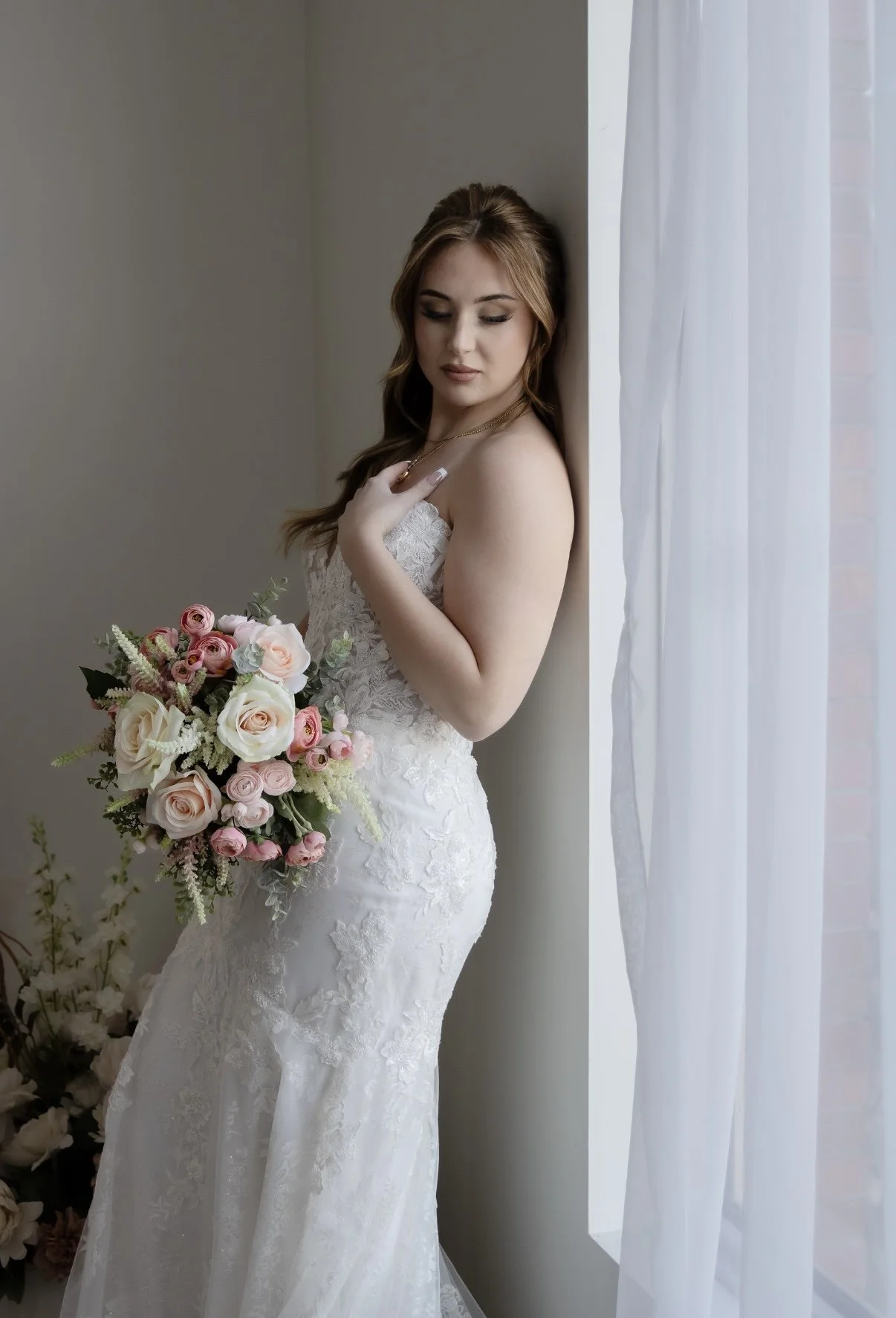 A bride in a white lace wedding dress holding a bouquet of pink and white roses, standing near a window with white curtains.