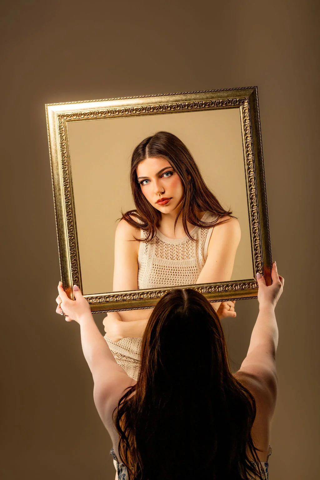 A young woman with brown hair and blue eyes looking into a large ornate picture frame held by another person's hands, with a plain beige background.