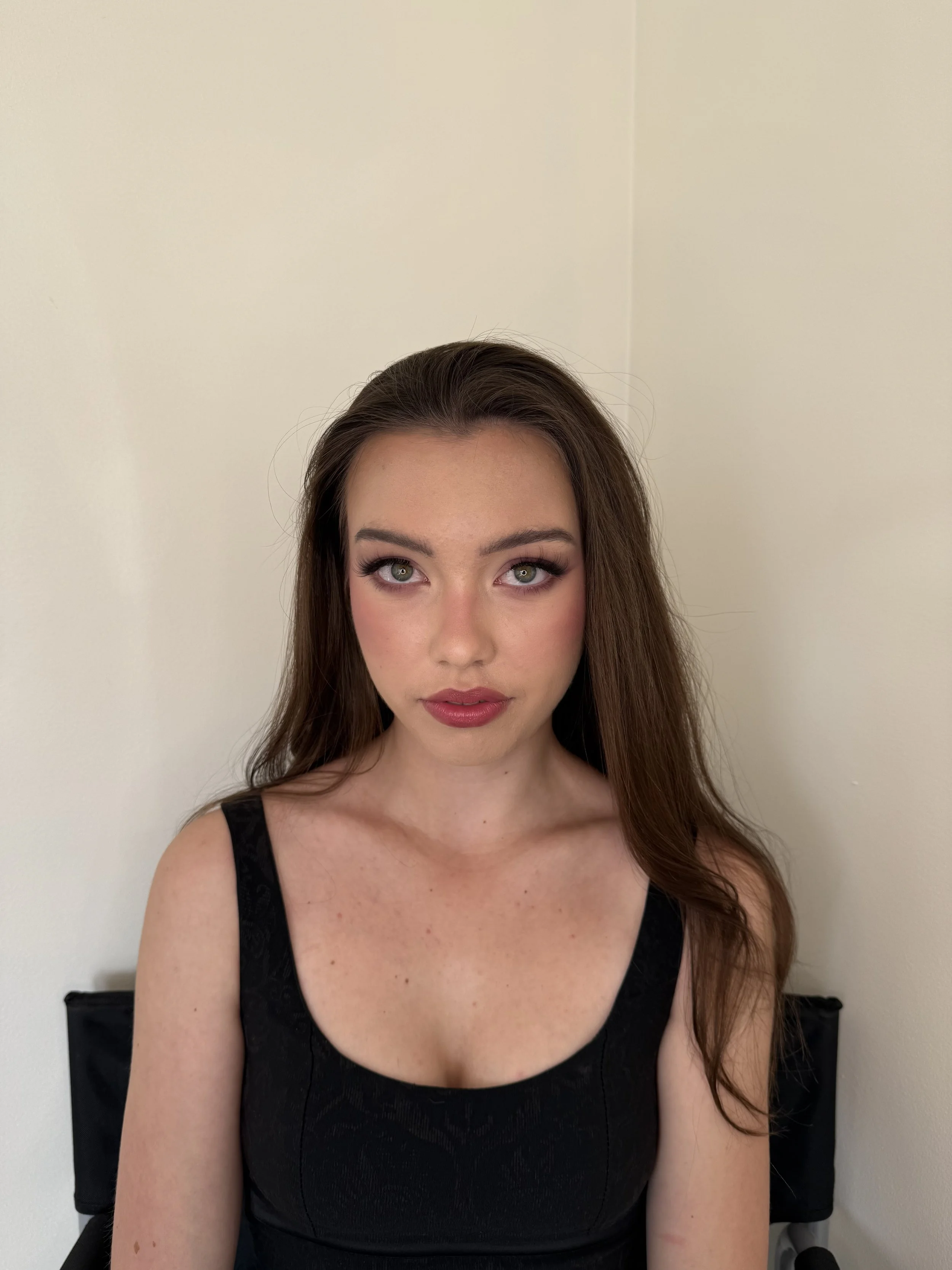 A woman with long brown hair and green eyes wearing a black sleeveless top, sitting in front of a plain beige wall.