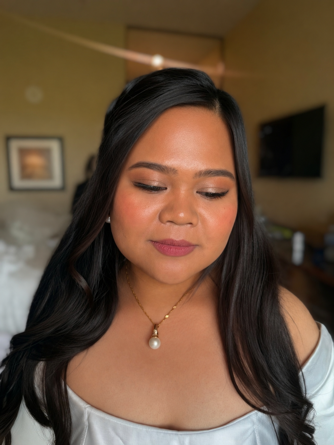 Close-up of a woman with long dark hair, wearing a white off-shoulder top, pearl necklace, and makeup, in a softly lit room.