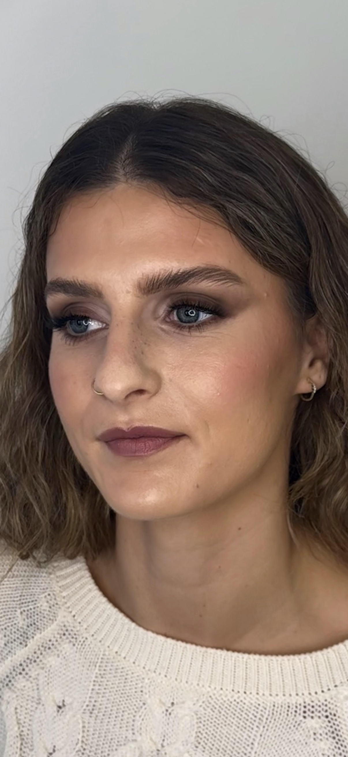 Close-up of a woman with wavy brown hair, blue eyes, wearing makeup, small hoop earrings, and a beige knit sweater.